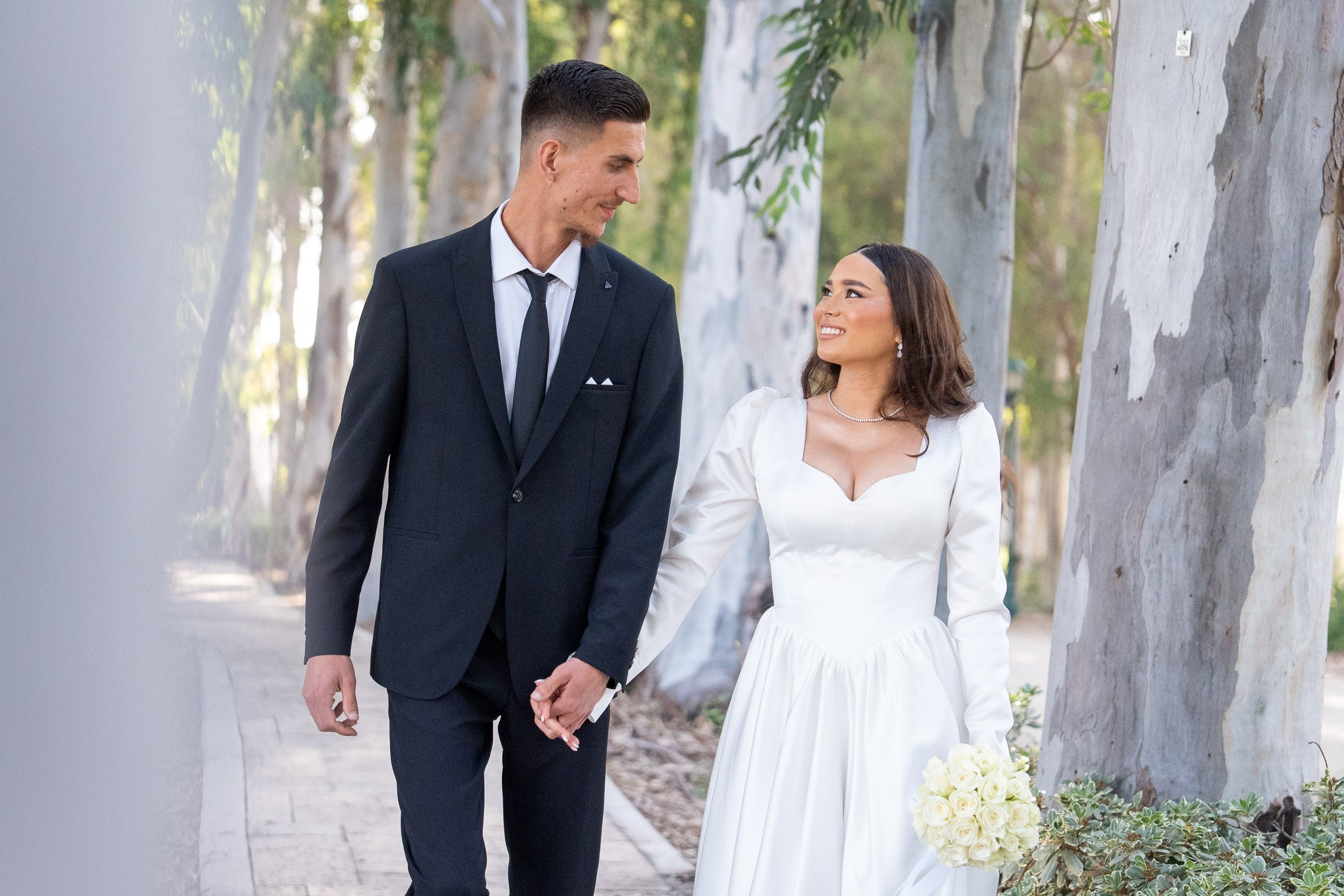 A bride and groom walk hand in hand along a pathway lined with eucalyptus trees