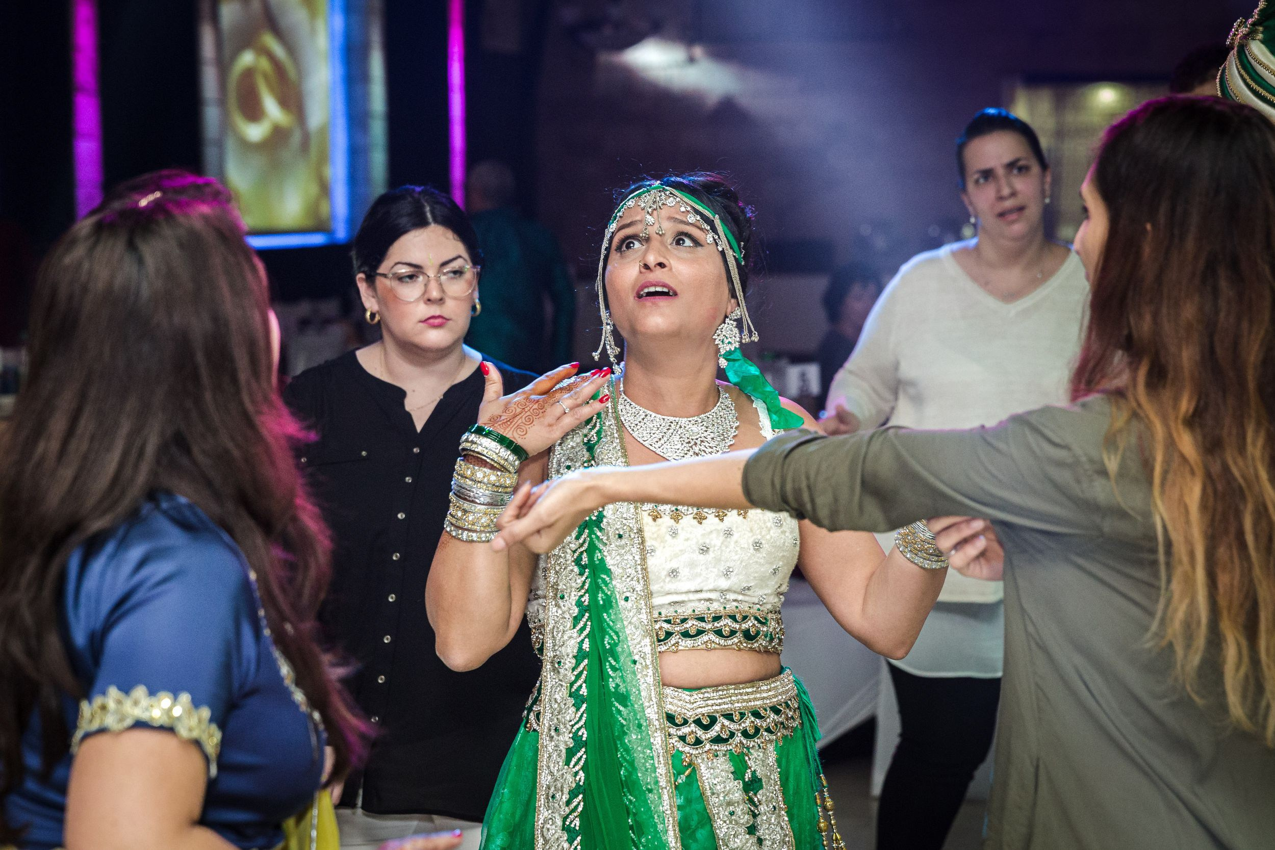 A bride in a green and white traditional Indian outfit is surrounded by guests on the dance floor. She has an emotional expression, adorned with intricate jewelry, bangles, and henna designs on her hands. The background features dramatic lighting with guests reacting to the moment, some appearing surprised or engaged in conversation. The image captures the energy and emotions of a wedding celebration.