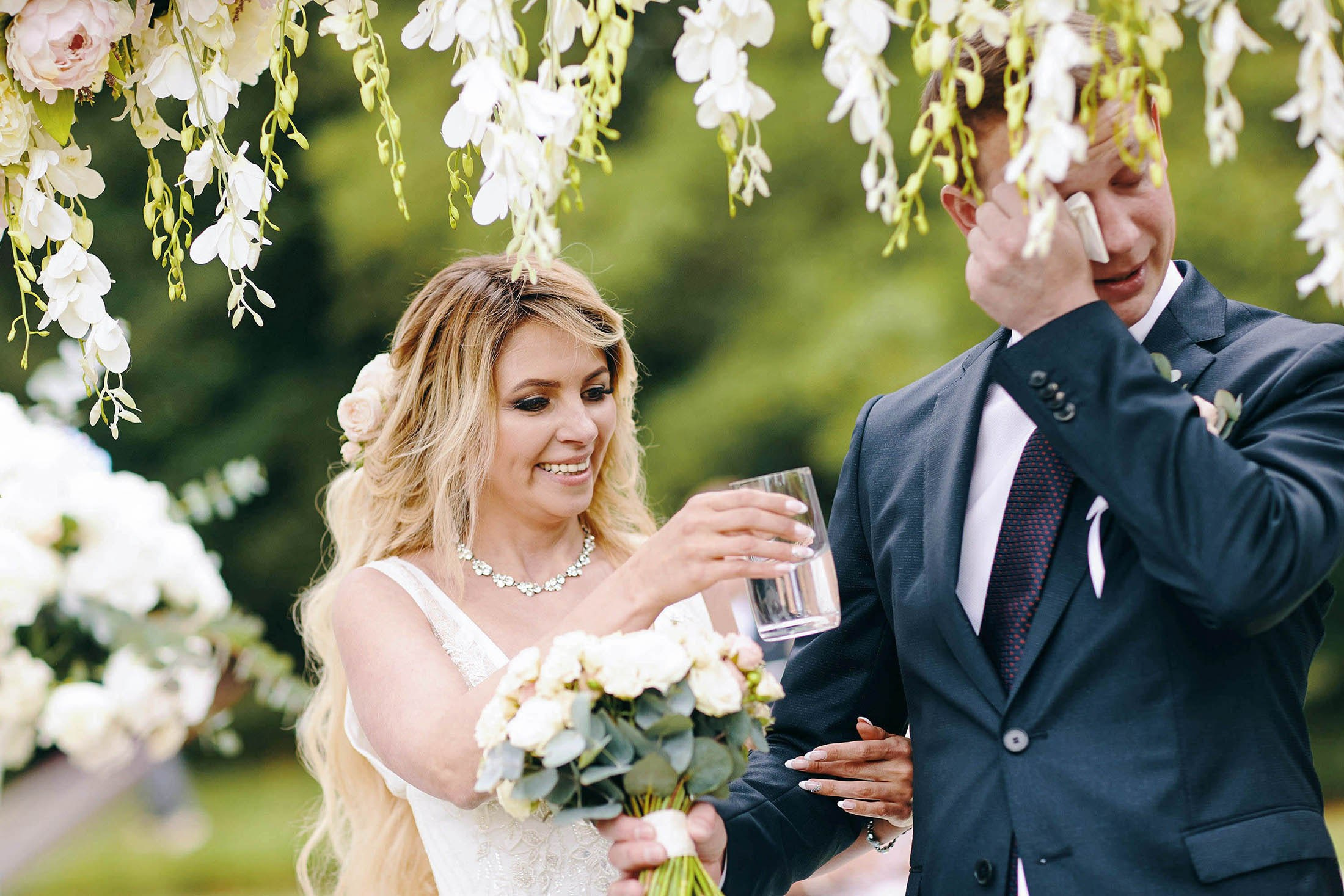 Bride handing water to crying groom under floral arch, Castle Hluboká ceremony