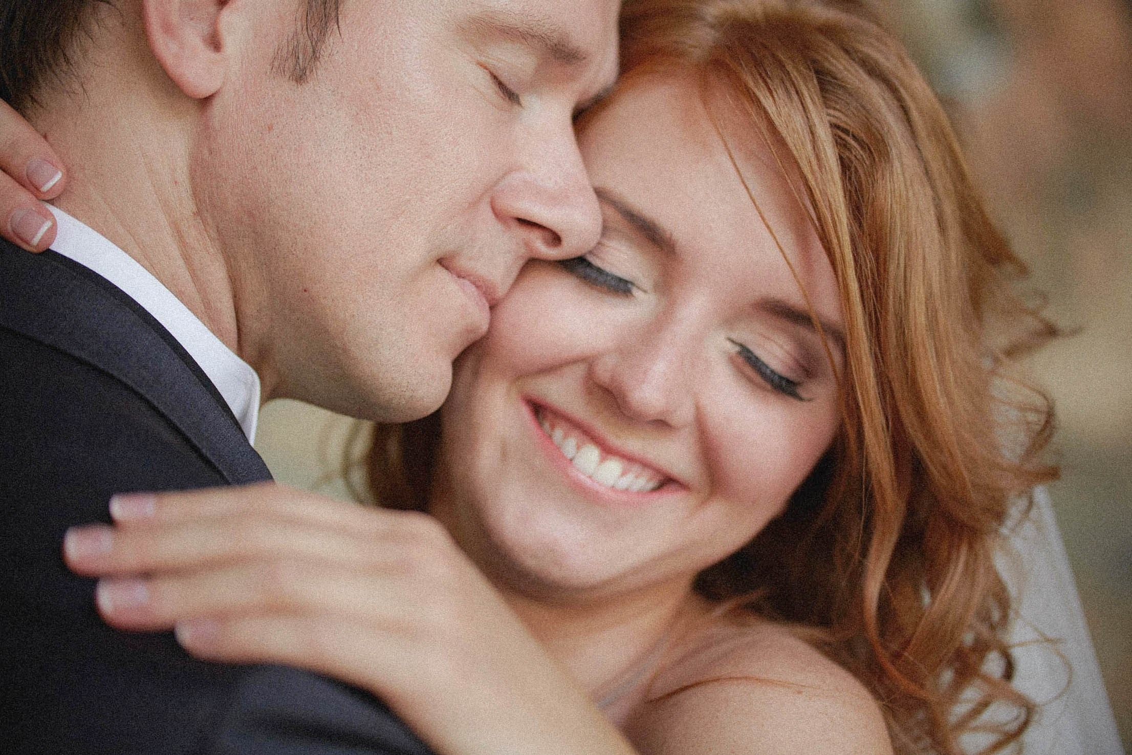 Smiling, red-headed bride, lovingly embraced by groom following their wedding ceremony at the Vrtba Garden.
