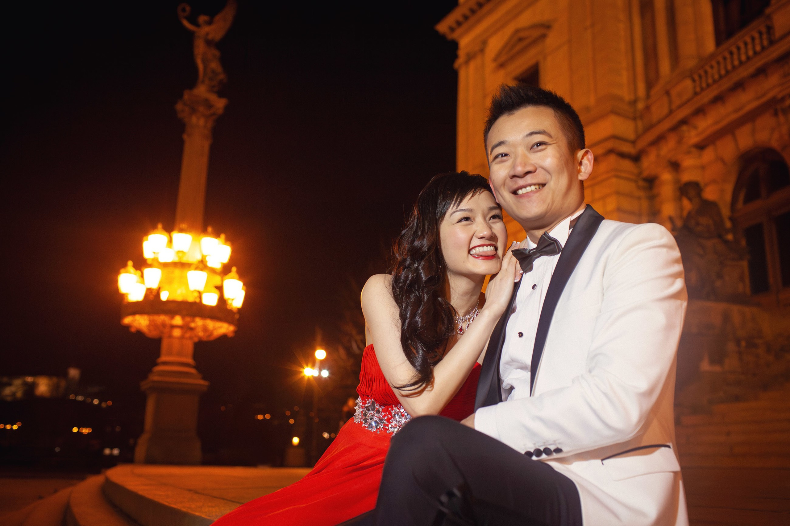 Beaming Asian woman wearing a red evening dress with her tongue out smiles lovingly towards her white tuxedo-clad partner as they sit atop the steps of the Rudolfinum in Prague at night