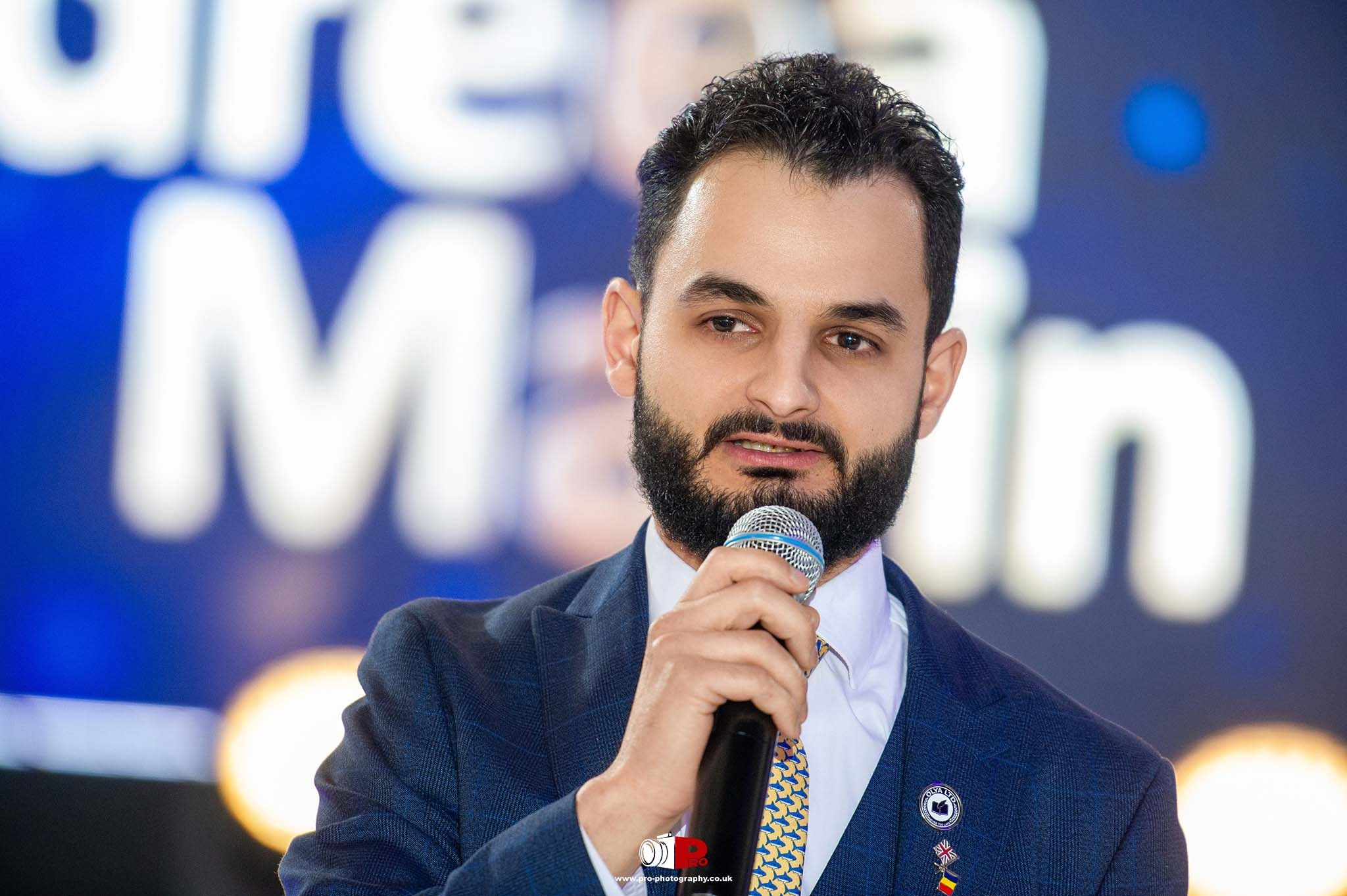 A bearded man in a blue suit is speaking passionately into a microphone at a gala event.