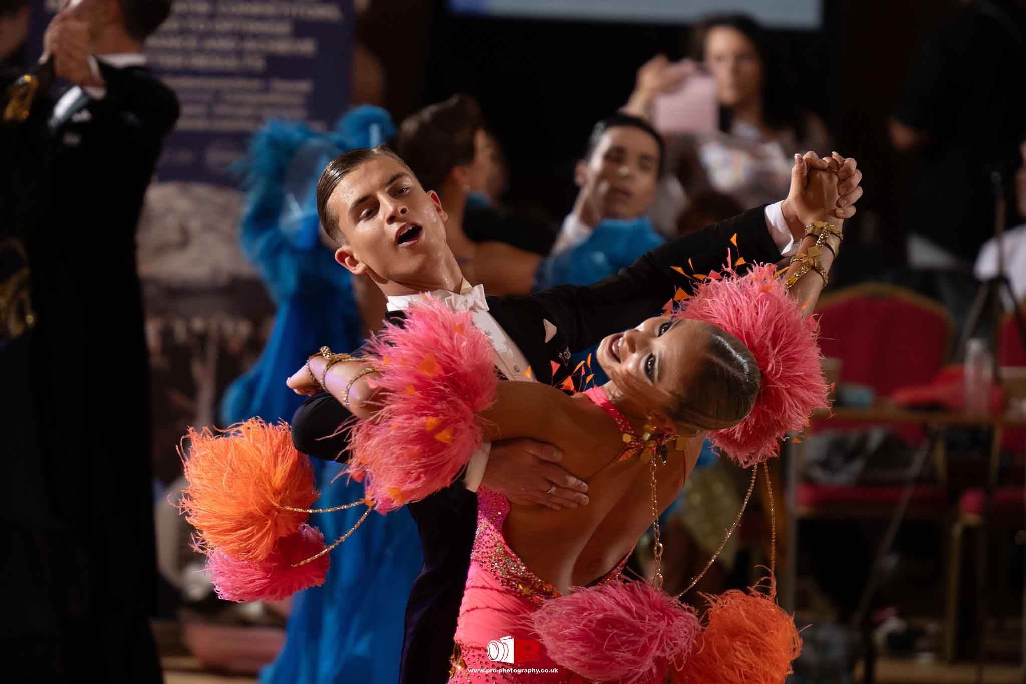 Professional ballroom dance couple performing a synchronized routine in vibrant pink and orange feathered costumes.