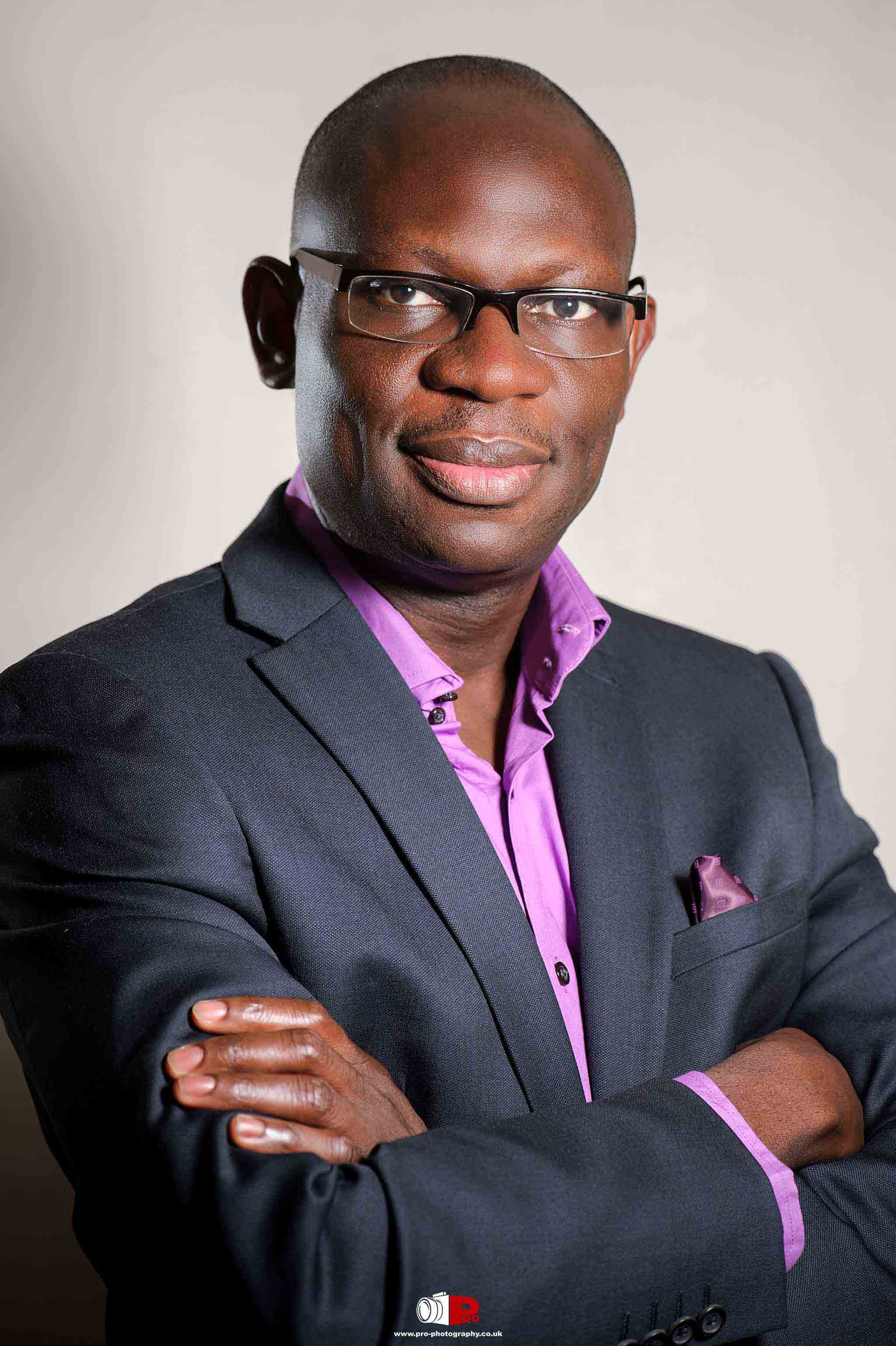 Professional portrait of a confident African-American businessman in a black suit with a purple shirt and pocket square, posing with crossed arms.