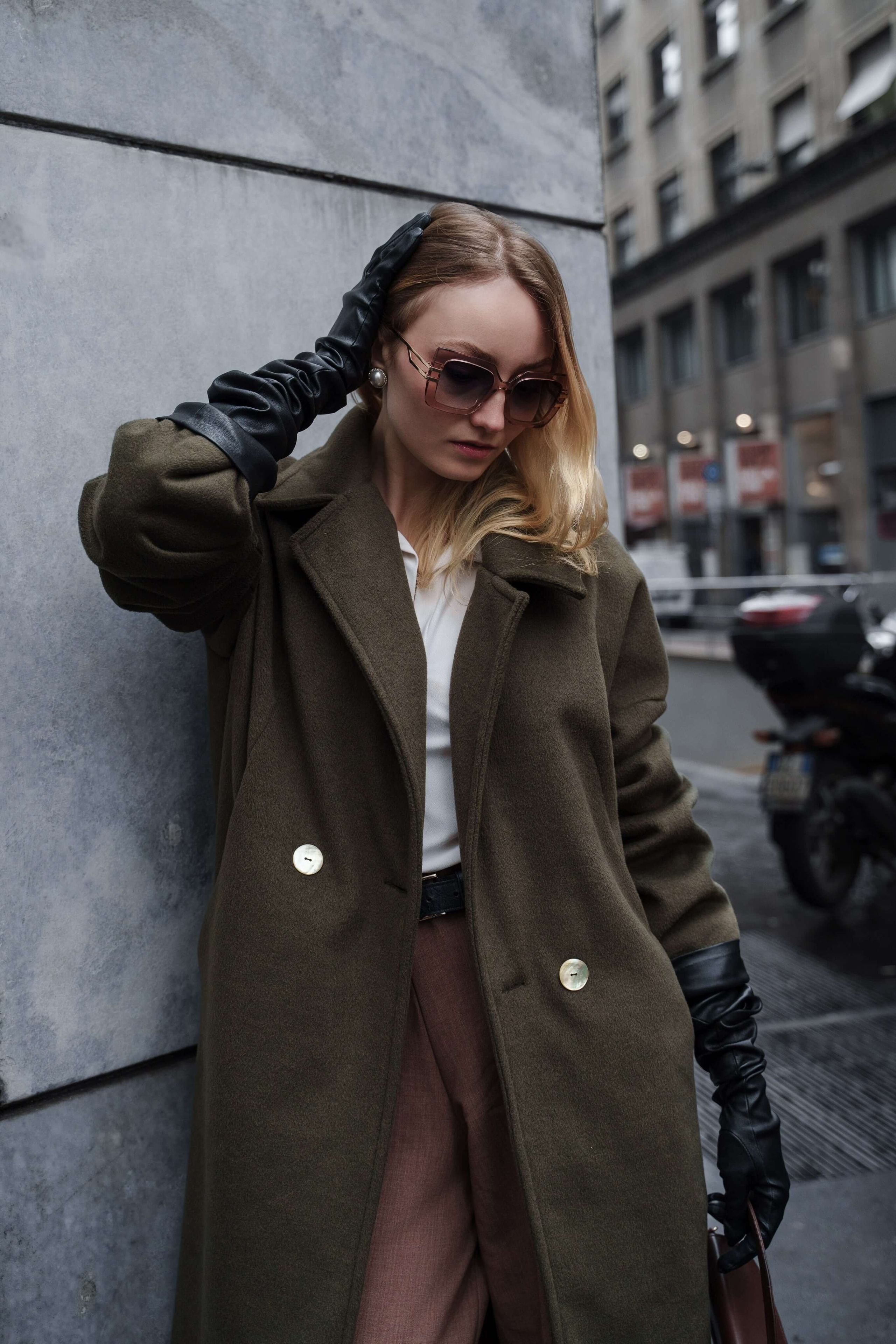 Elegant woman in sunglasses and long gloves posing with hand on head by a grey stone wall