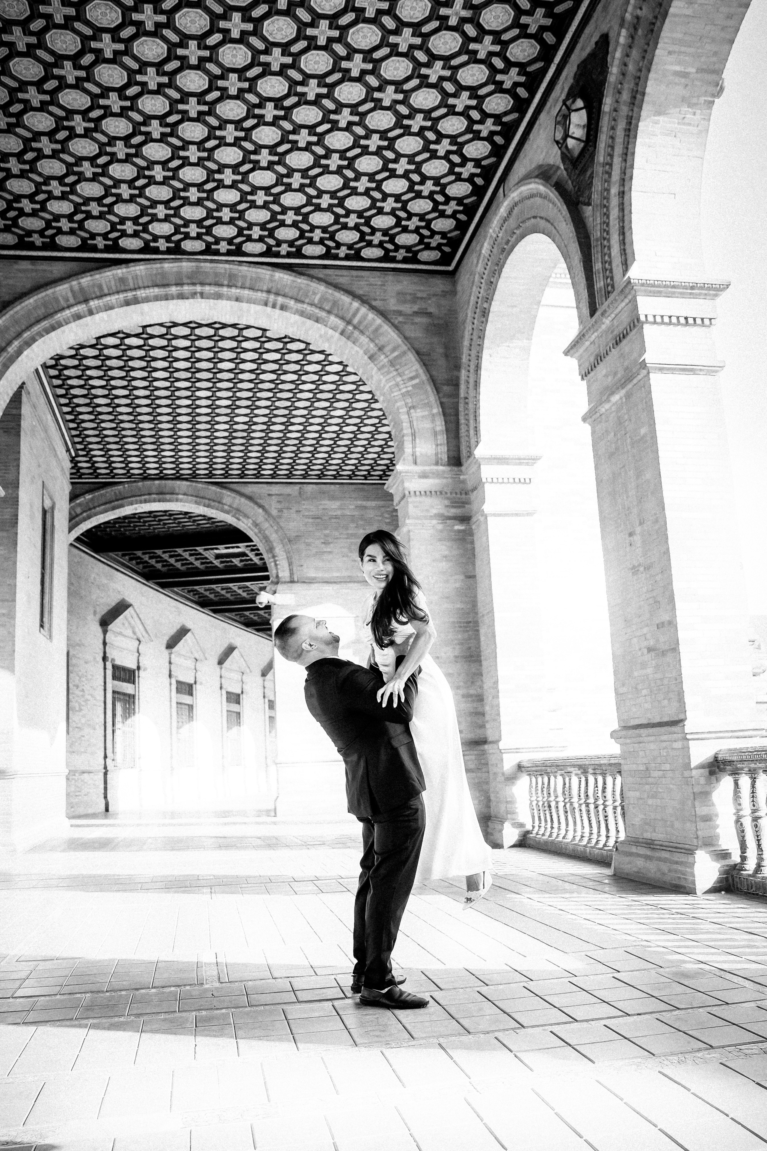 Black and white portrait of the groom lifting the bride beneath the arches of Plaza de España in Sevilla. Artistic intimate wedding photo highlighting the elegant architecture and romantic movement.
