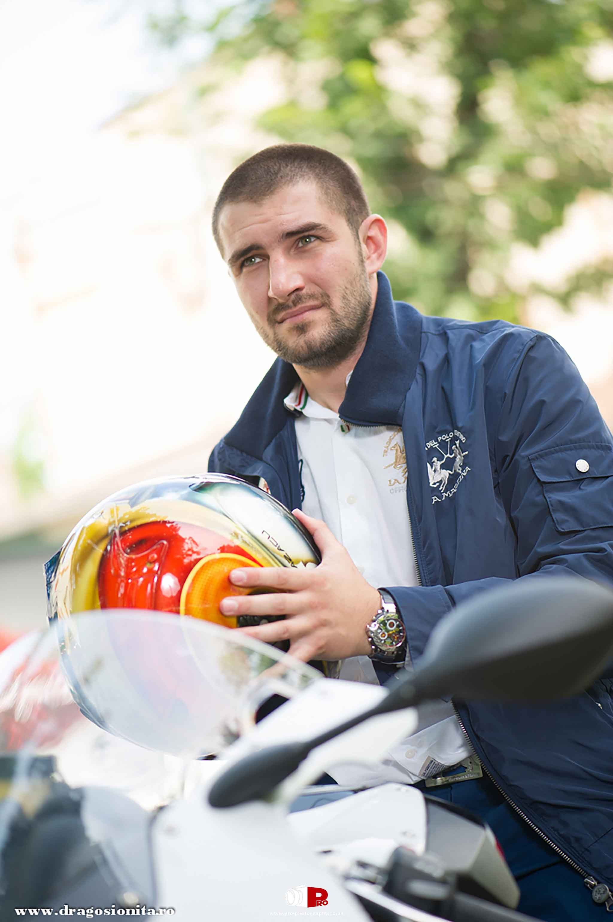A bearded man in a navy jacket holding a colourful helmet next to his motorcycle.