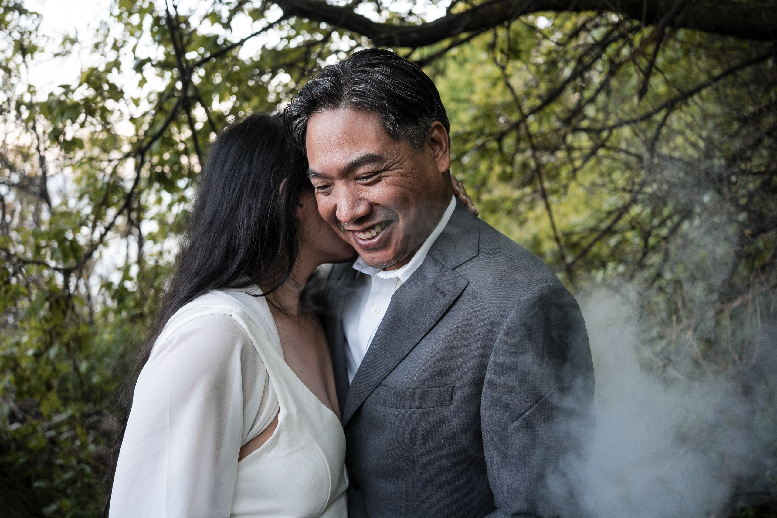 Engaged couple sharing an intimate and joyful moment in the forest, as the bride-to-be whispers in her fiancé’s ear, surrounded by soft smoke and evening light.