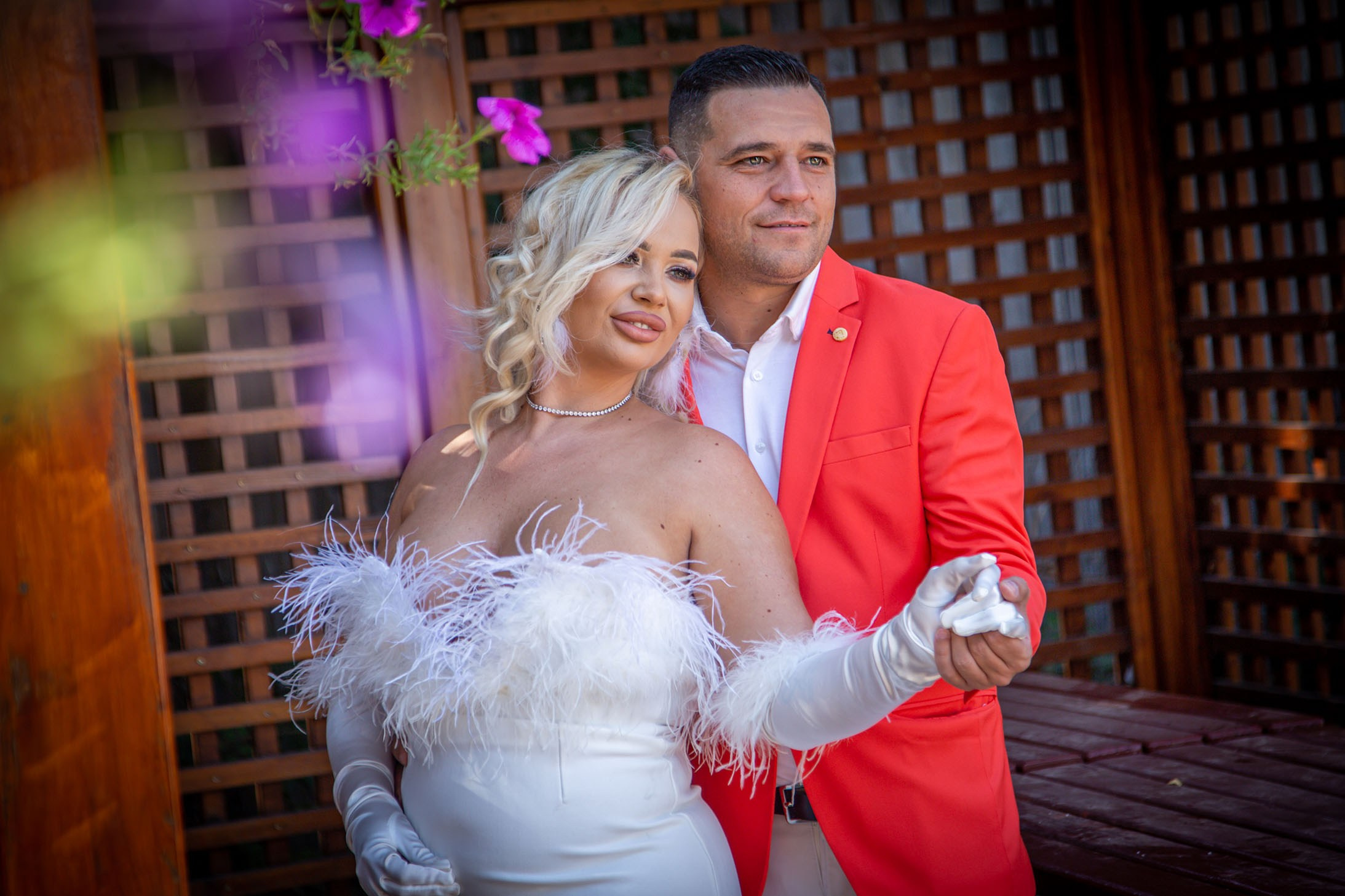 Mireasă blondă în rochie albă sta cu capul rezemat pe mire în sacou roșu, ședință foto romantică în aer liber 