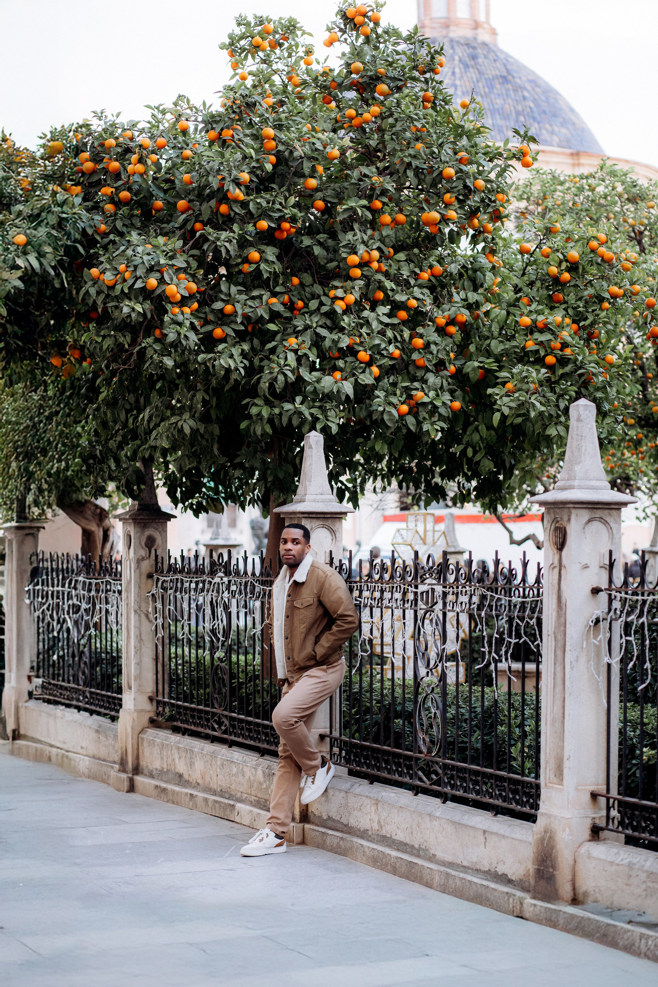 Мужская портретная фотосессия в Валенсии, Испания, со стильным мужчиной, позирующим у исторического кованого забора на фоне пышных апельсиновых деревьев с созревшими плодами, снятым при мягком естественном дневном свете с расслабленной средиземноморской атмосферой — идеально для мужской портретной фотографии, lifestyle-фотосессий для мужчин, городских outdoor-портретов, fashion-вдохновлённых съёмок и профессиональных мужских фотосессий в Валенсии и по всей Испании.
