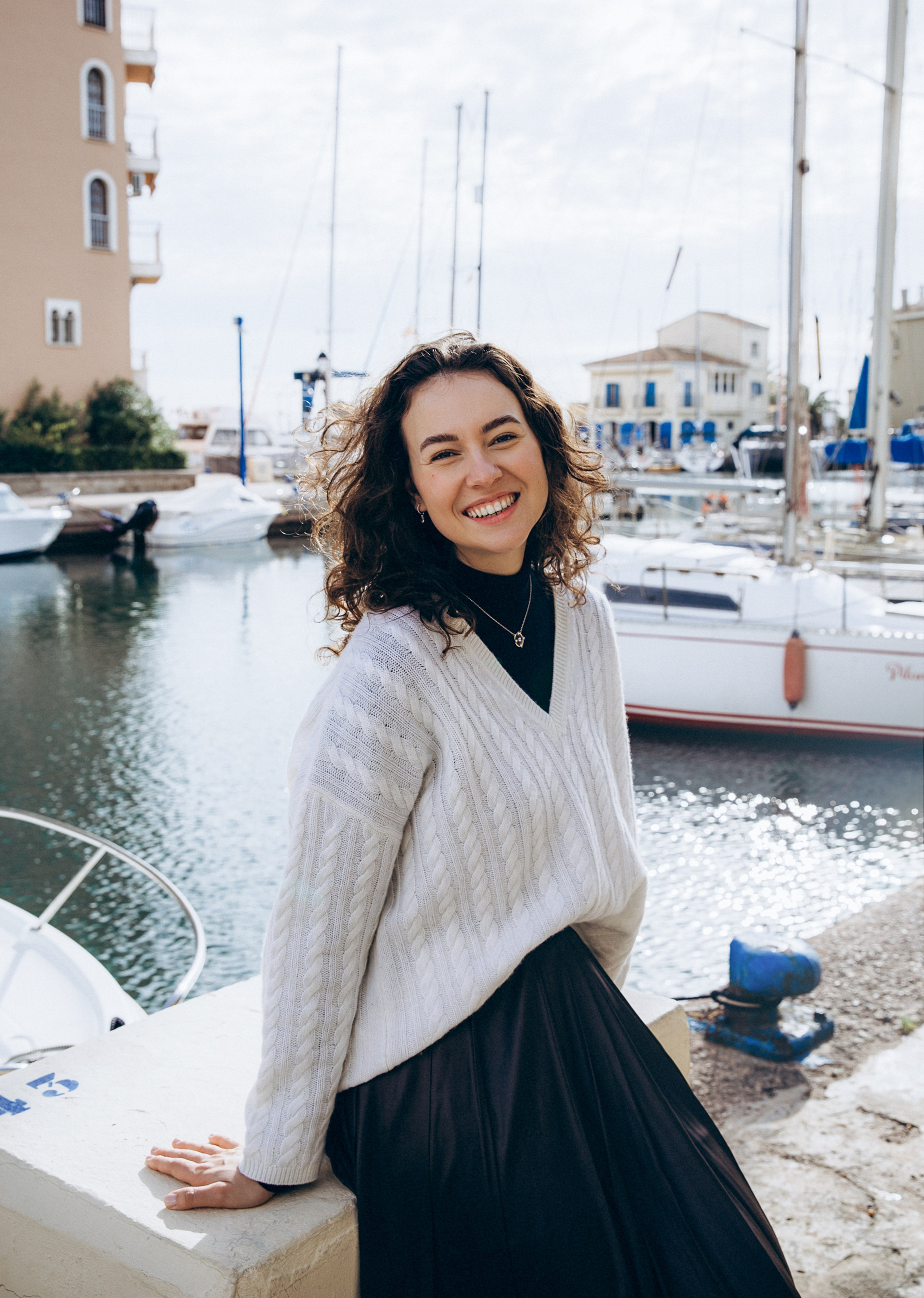 Женская фотосессия в Валенсии, Испания, с улыбающейся женщиной с естественными кудрями у марины, с парусниками и спокойной водой на фоне, снятая в мягком дневном свете с расслабленным и искренним настроением — идеально для женской фотографии, портретных сессий, лайфстайл-съёмок, живых портретов и профессиональных женских фотосессий в Валенсии и по всей Испании.