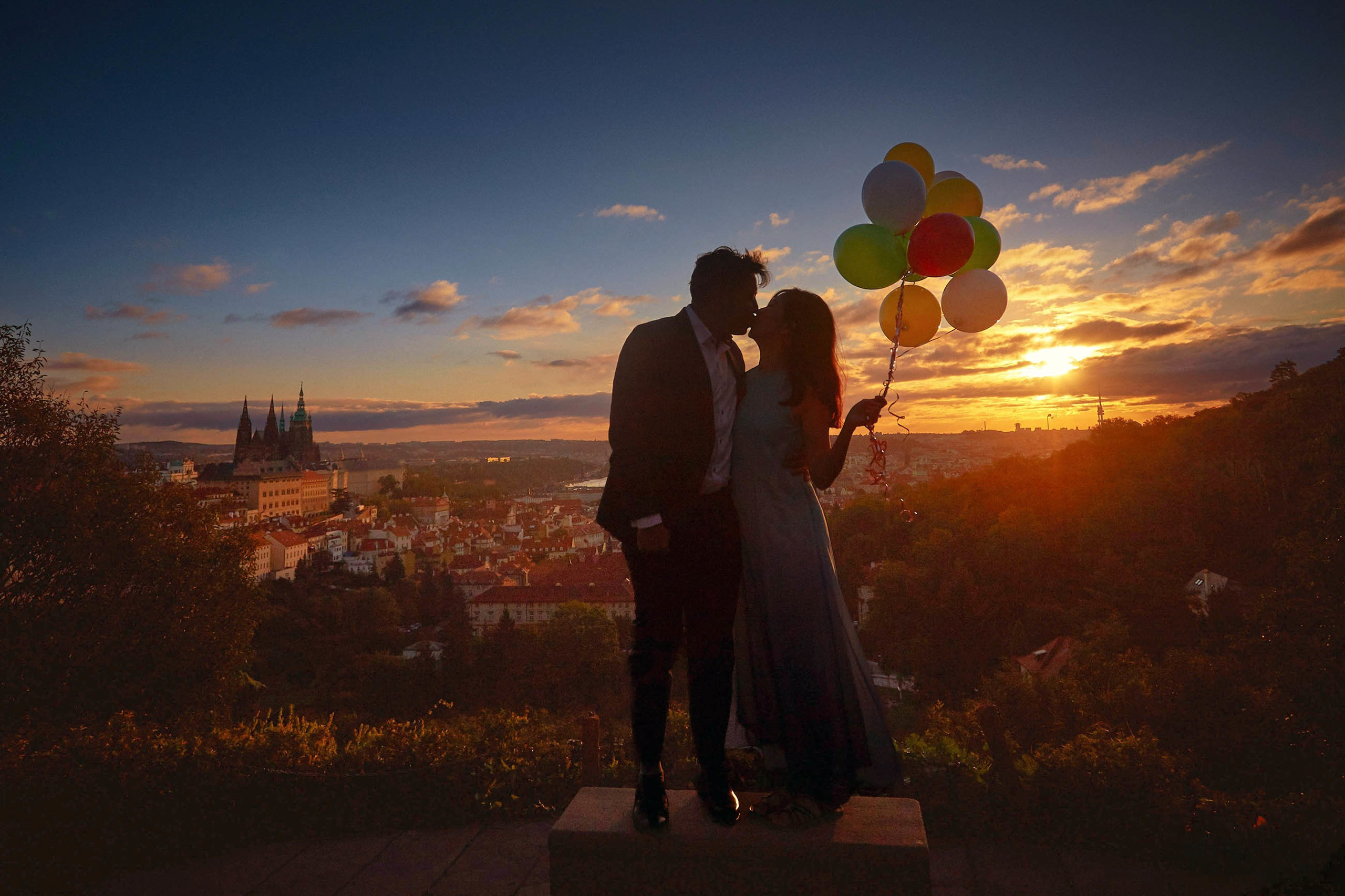 Silhouetted couple kissing with balloons against a rising sun and Prague skyline in the background.