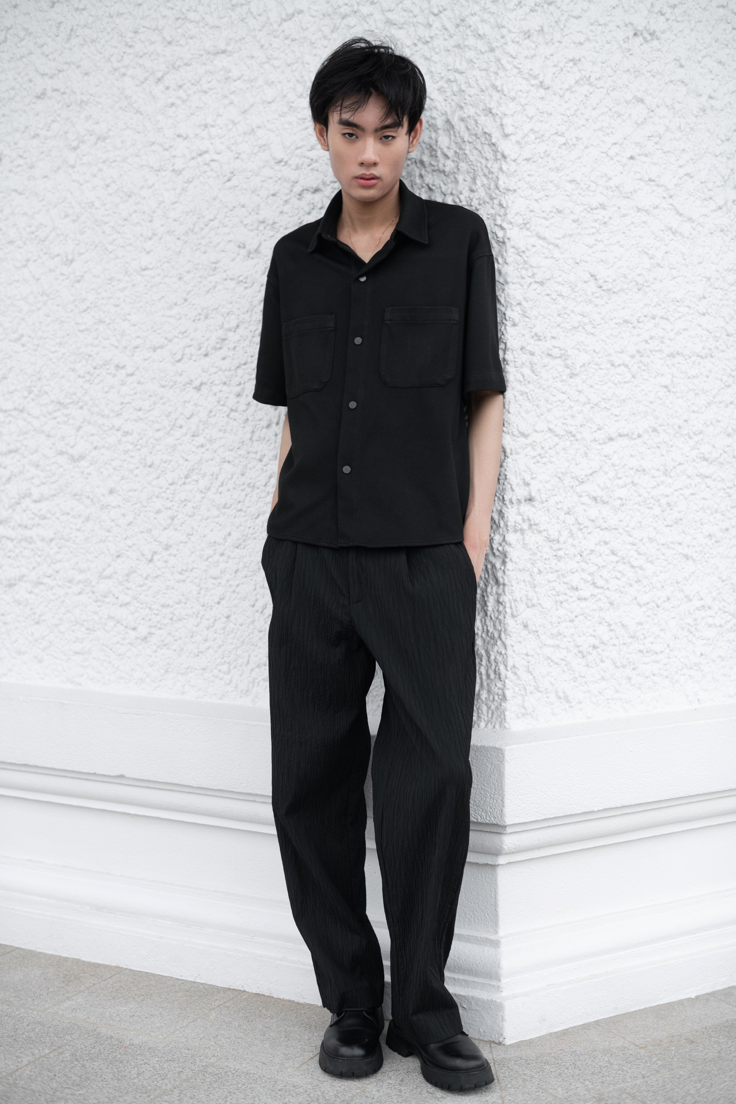 Male model in black outfit posing against a white textured wall in Singapore fashion editorial.