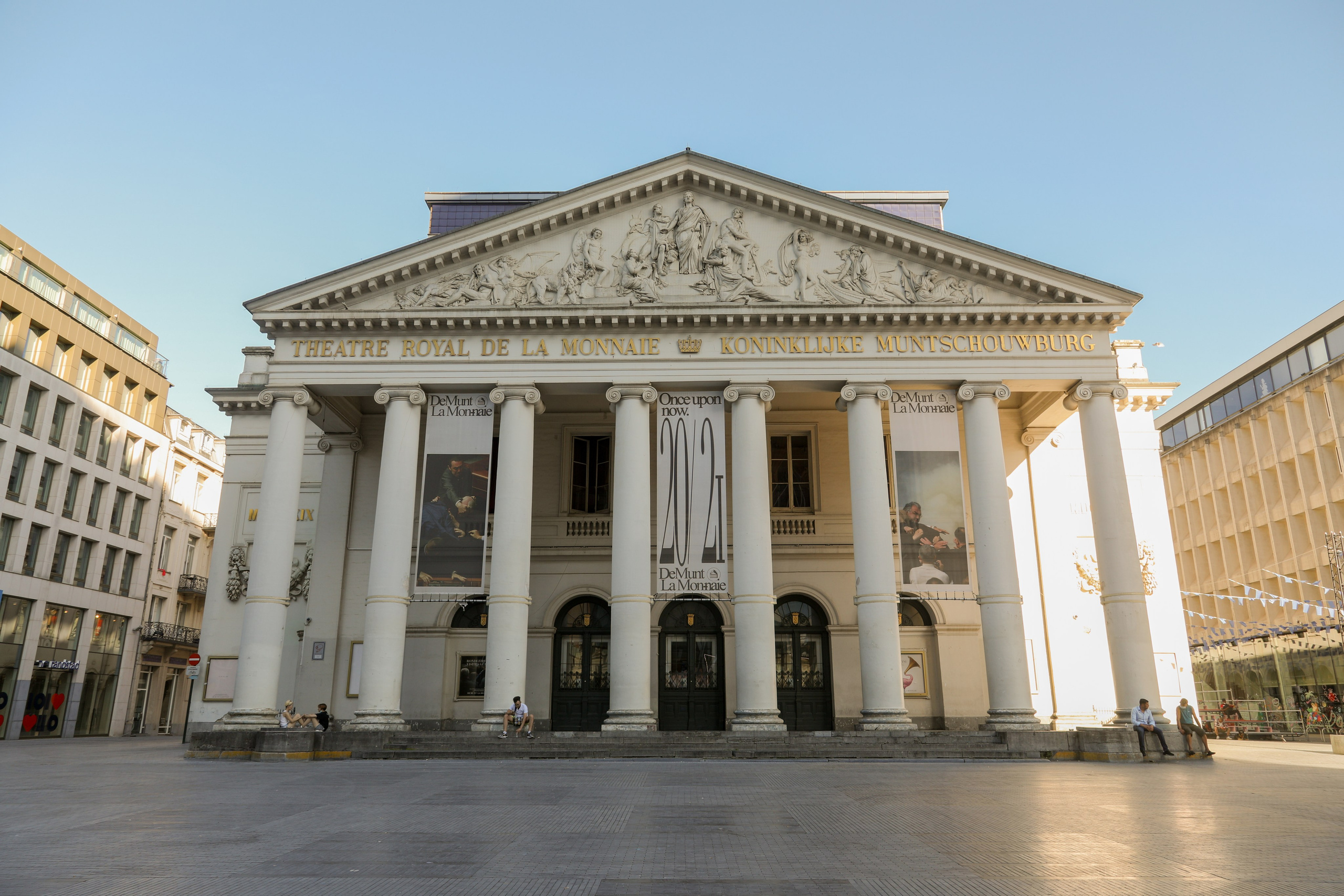 A wide shot of the grand, white neoclassical Royal Theatre of La Monnaie opera house in Brussels, featuring large columns, a detailed pediment, and street banners.