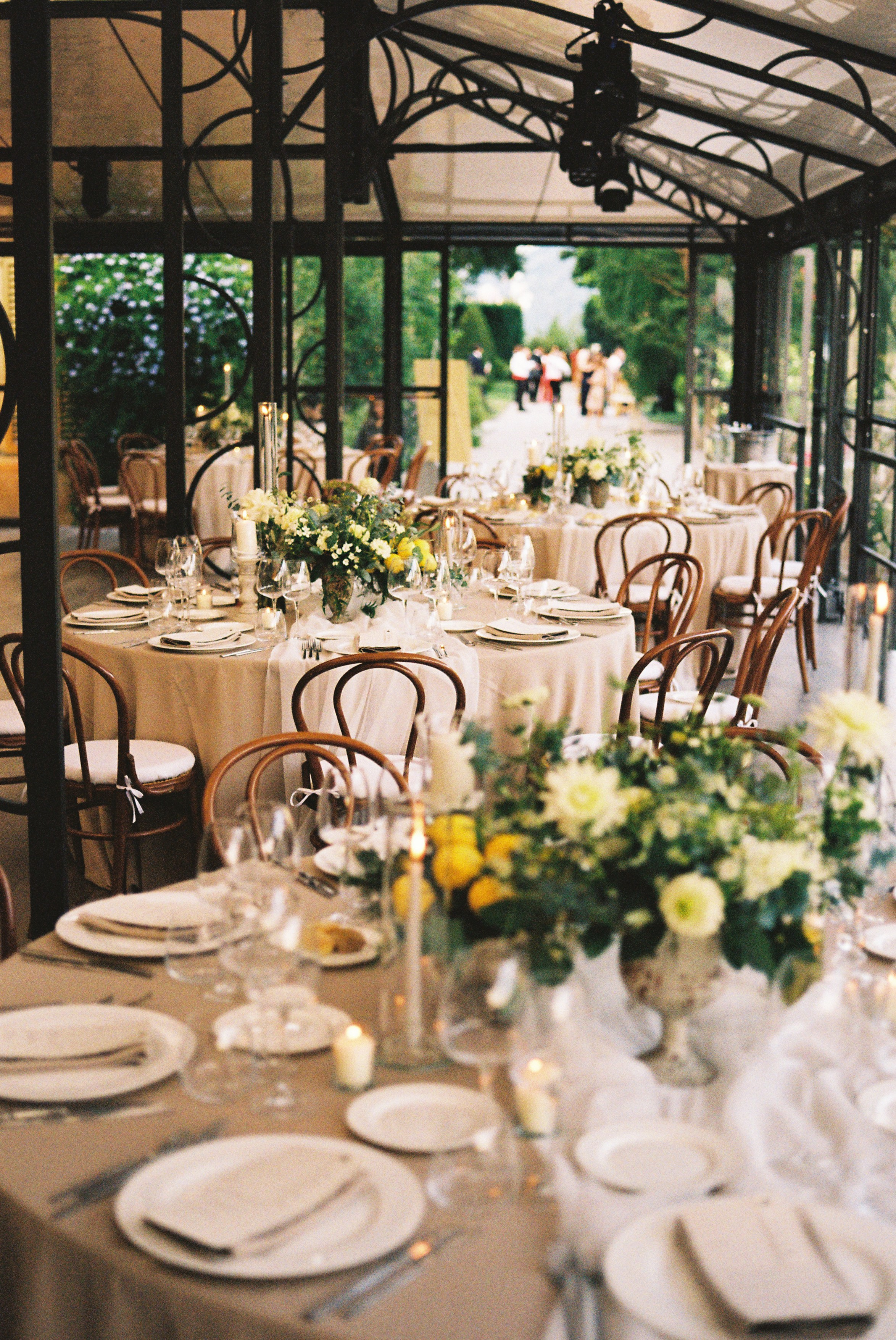 Elegant indoor wedding reception setup with round tables, floral centerpieces, and candlelight in glass pavilion.