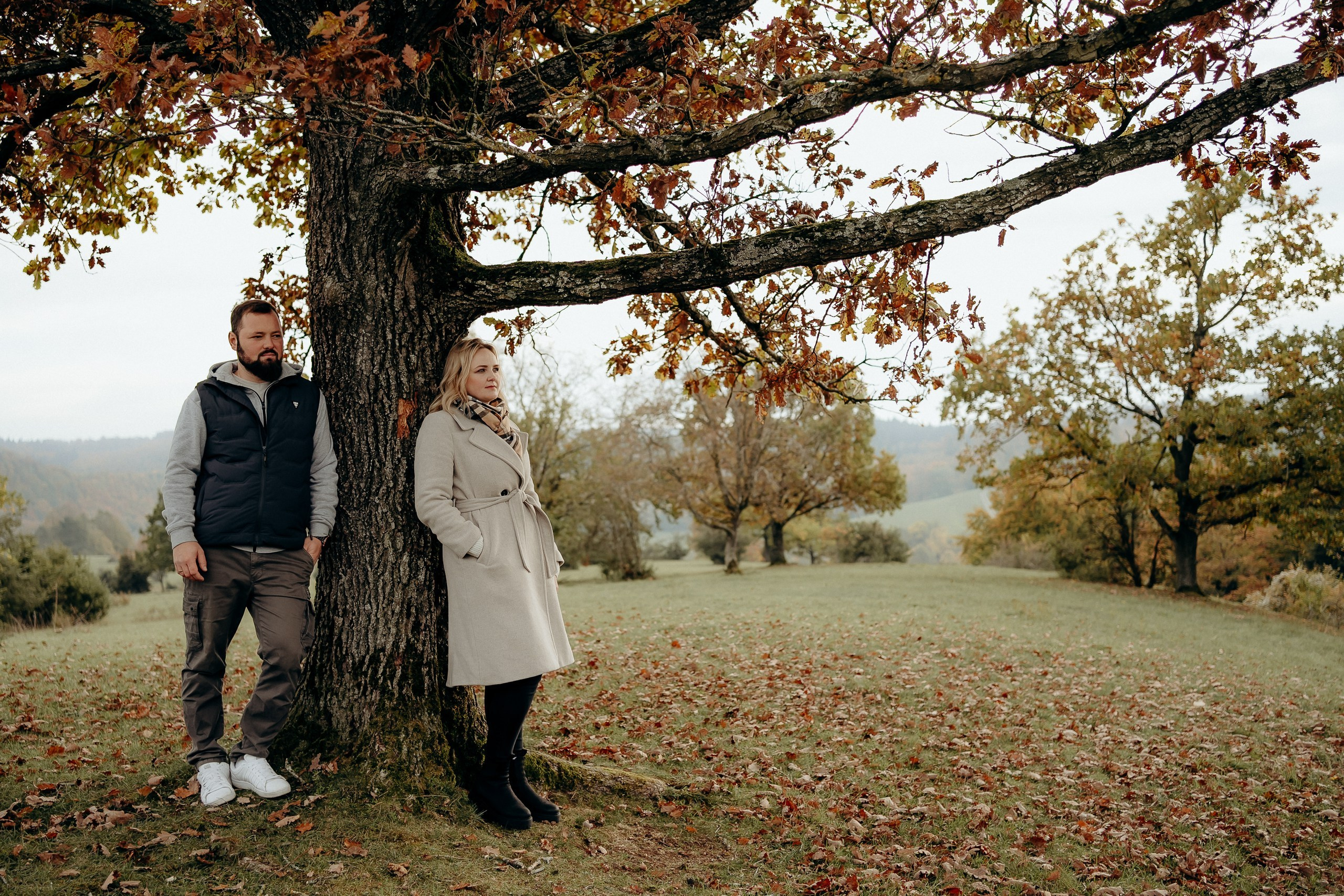 Paar steht gemeinsam unter einem großen Baum in einer herbstlichen Landschaft.