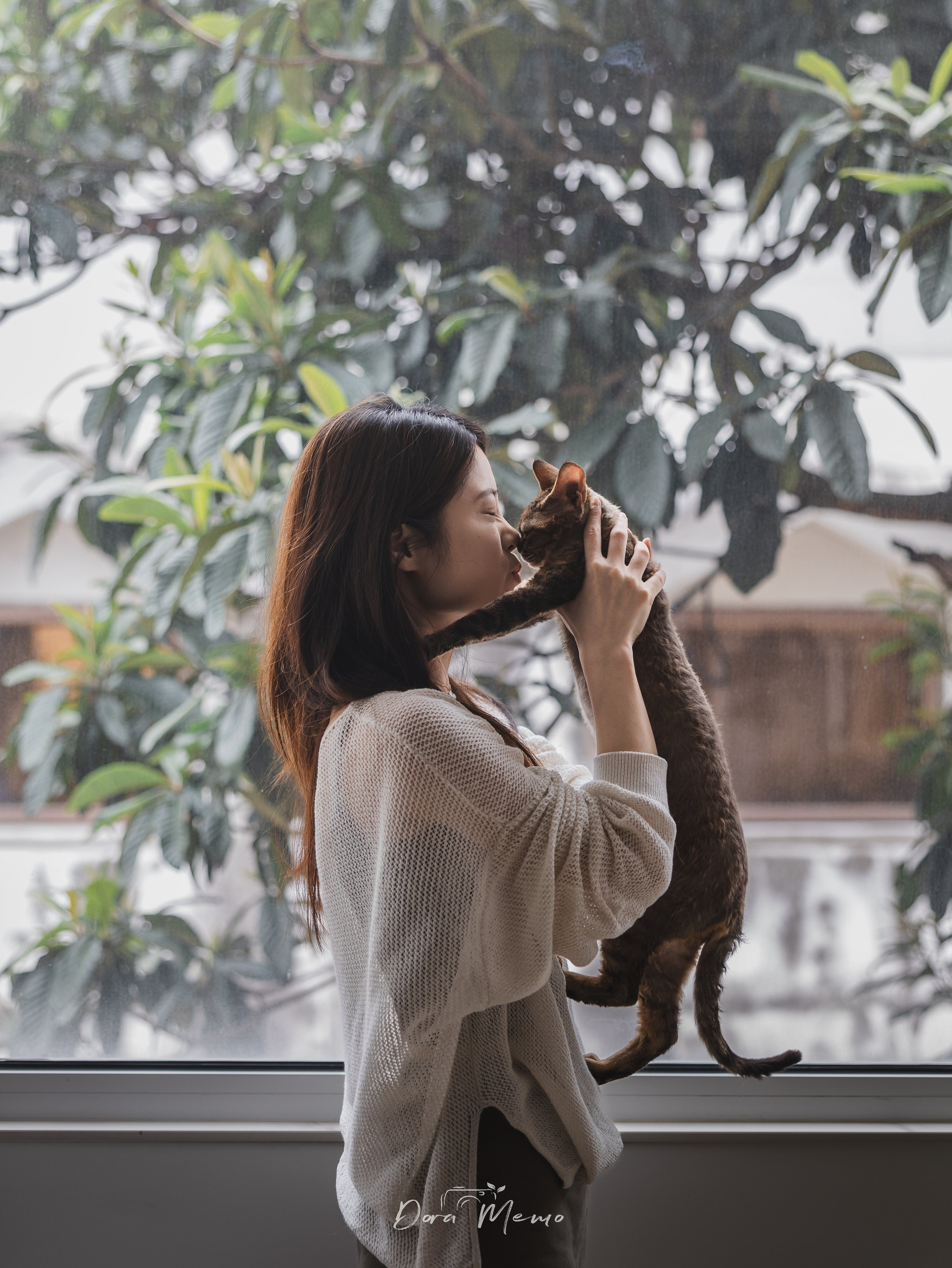 A woman gently kisses her cat by a large window during an at-home pet family photography session in Shanghai.