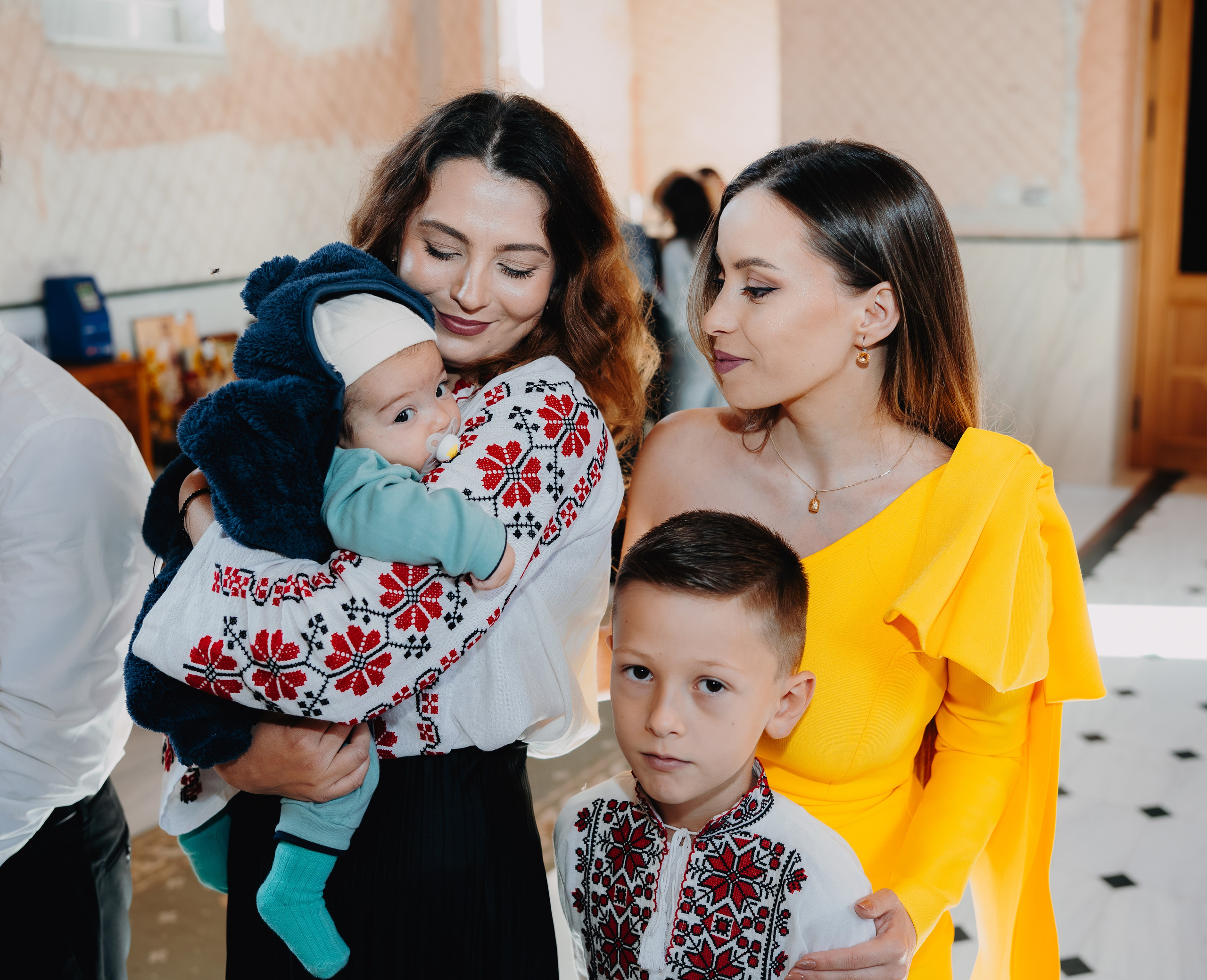 Close-up intim: Arthur Mihail, înfășat, este ținut cu grijă în brațele nașei. Nașul (fiul ei) stă protector lângă ei, cu o mână pe spatele nașei, într-un cadru plin de tandrețe și responsabilitate.