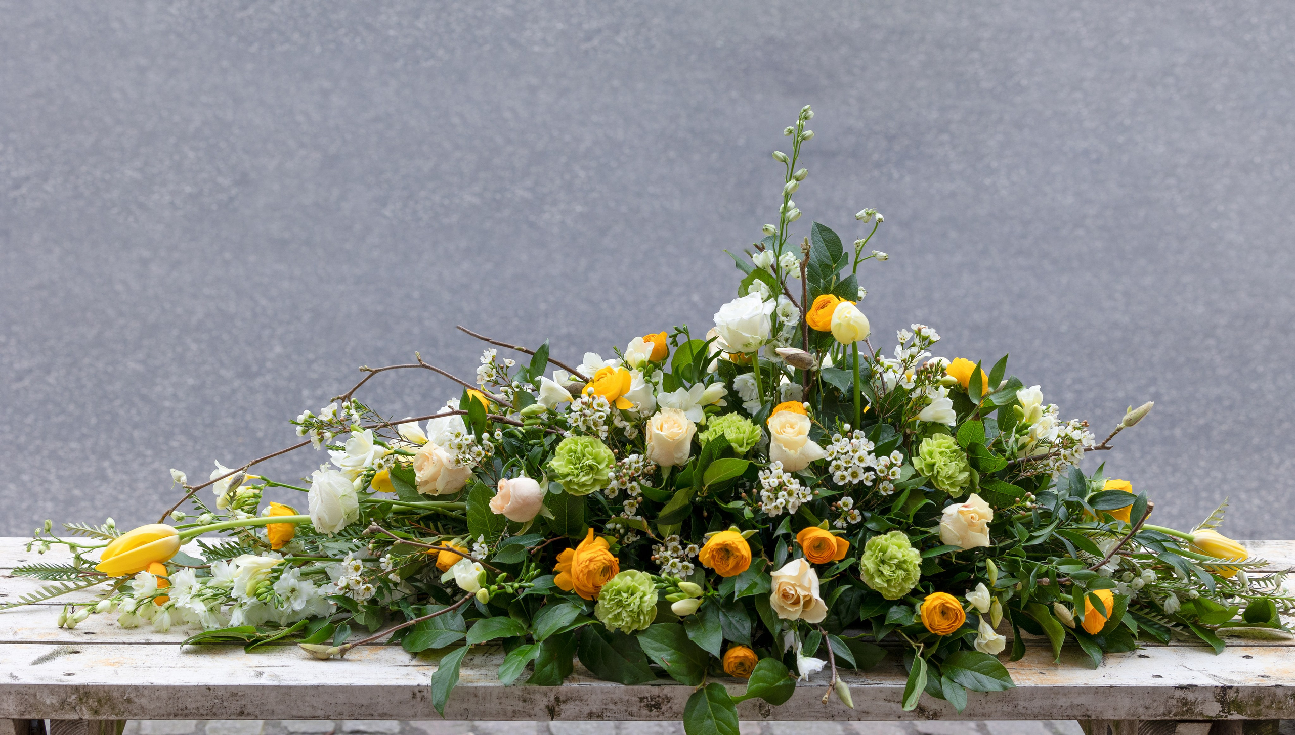 Blomsterne er arrangeret tæt på hele kistelåget, som giver en overdådig kiste. De flotte grønne, hvide og gule nuancer er kombineret smukt i sæsonens blomster. Et unikt farvel der tager sig elegant ud i kirkerummet.