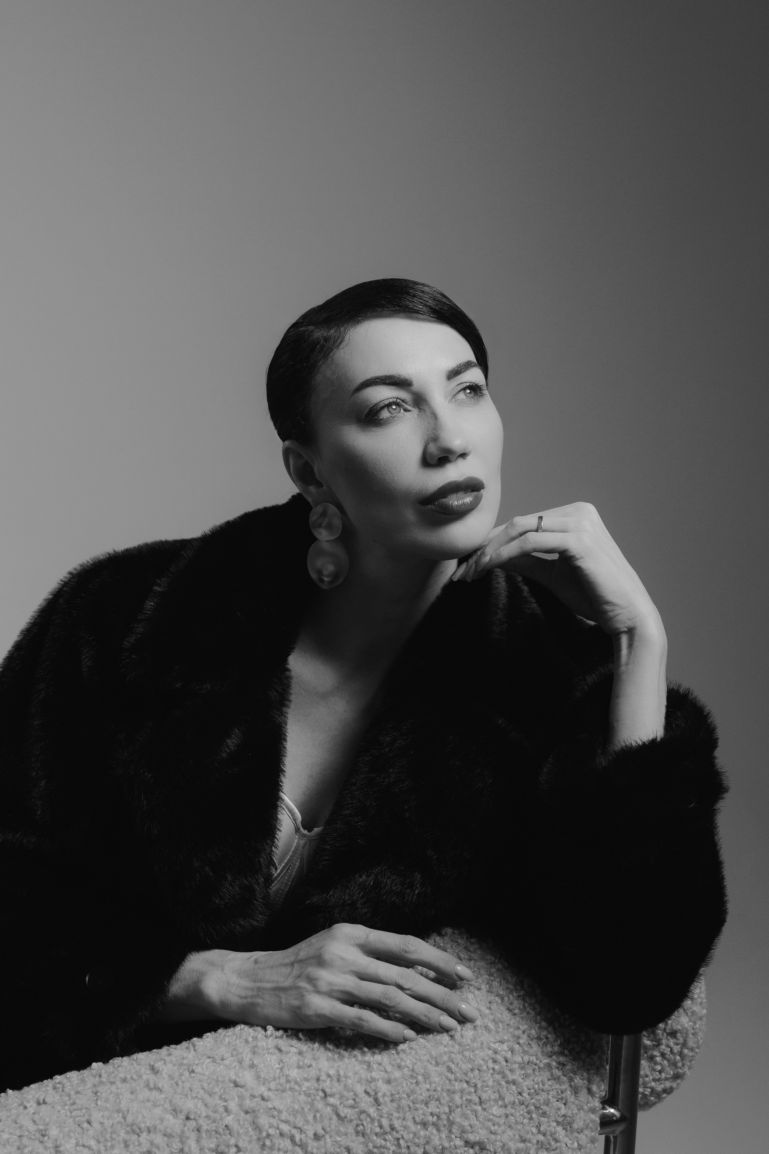 Black and white editorial portrait of a woman in a black fur coat sitting at a table with elegant pose, photographed in Leipzig, Germany. Черно-белый редакционный портрет девушки в чёрной шубе, сидящей за столом в элегантной позе, снятый в Лейпциге, Германия.