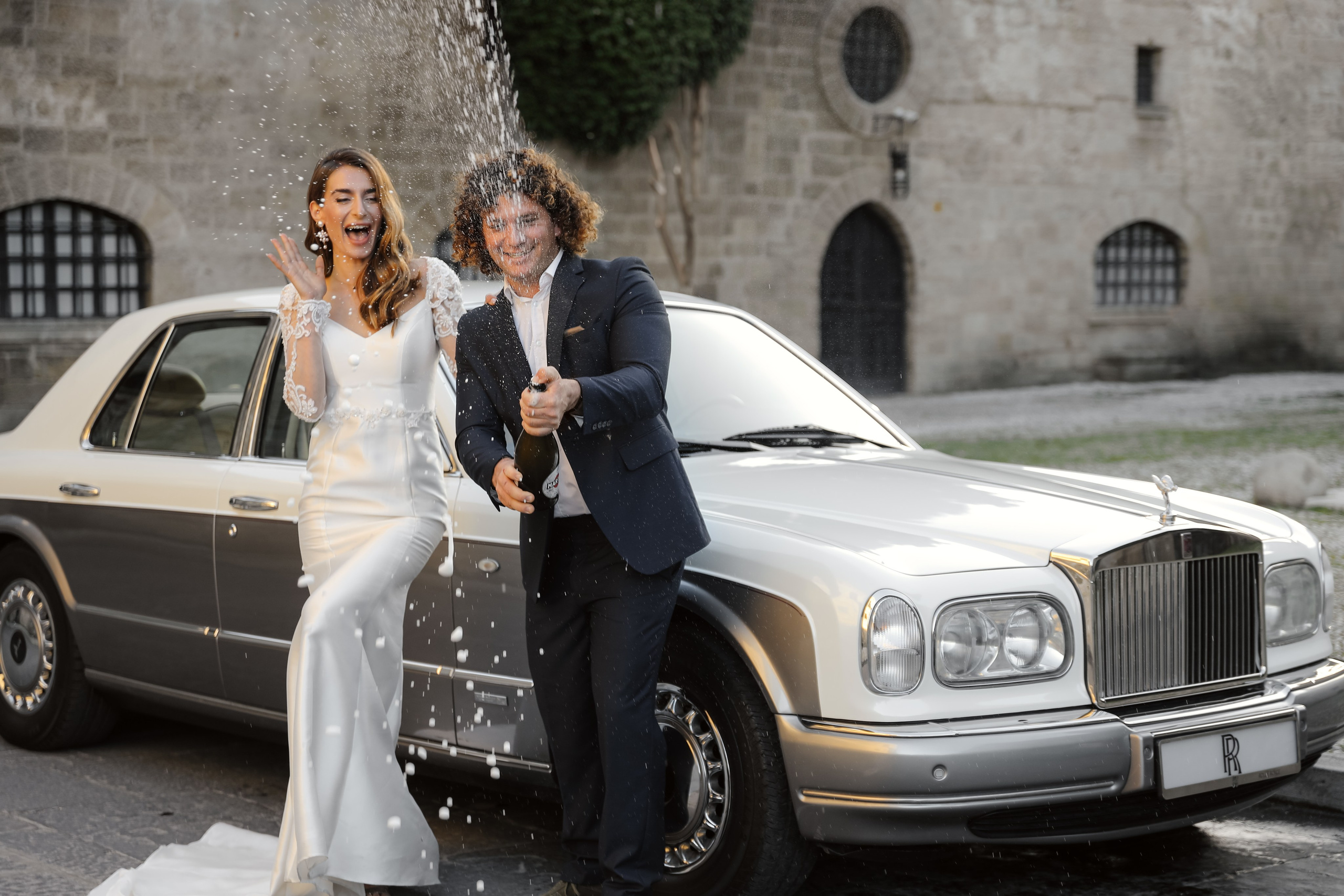 A bride and groom opening champaign next to Rolls Royce in old town of Rhodes island, Greece