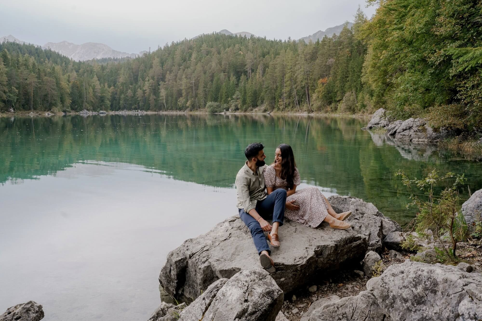 Couple sitting on rocks at Eibsee lake with turquoise water and mountains in background during engagement session