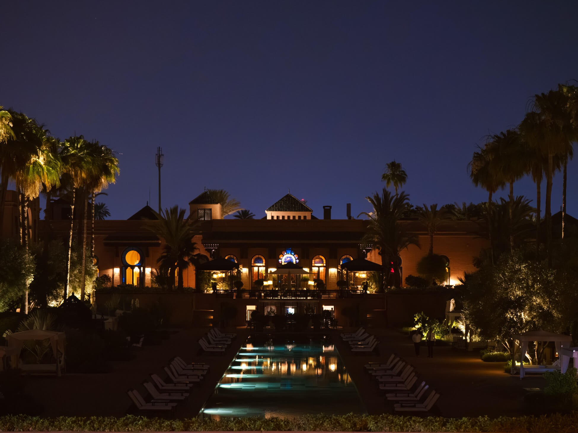 Luxury destination wedding venue in Marrakesh at night: the grand illuminated palace façade reflected in a long pool, palm silhouettes against a deep blue sky