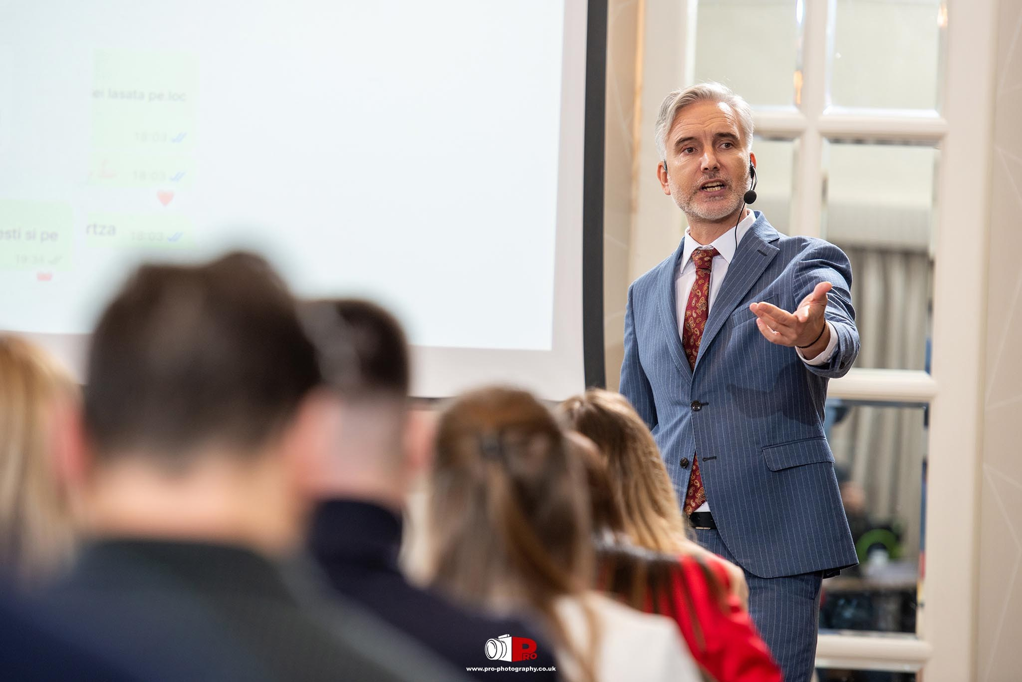 A well-dressed speaker delivering an engaging presentation with projected visuals to a focused audience.
