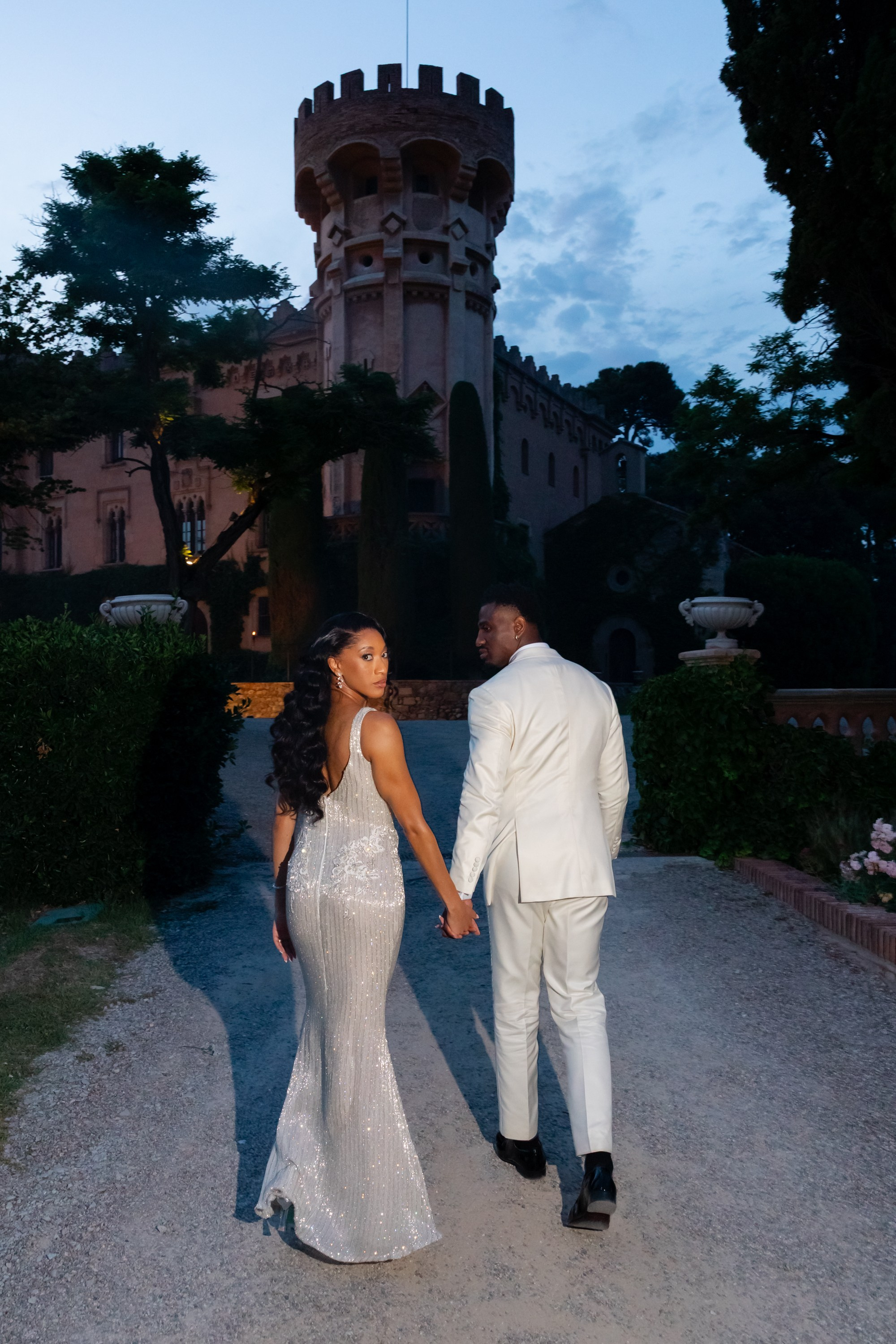 black groom and bride in a silver mermaid dress walking in twilight towards a castle