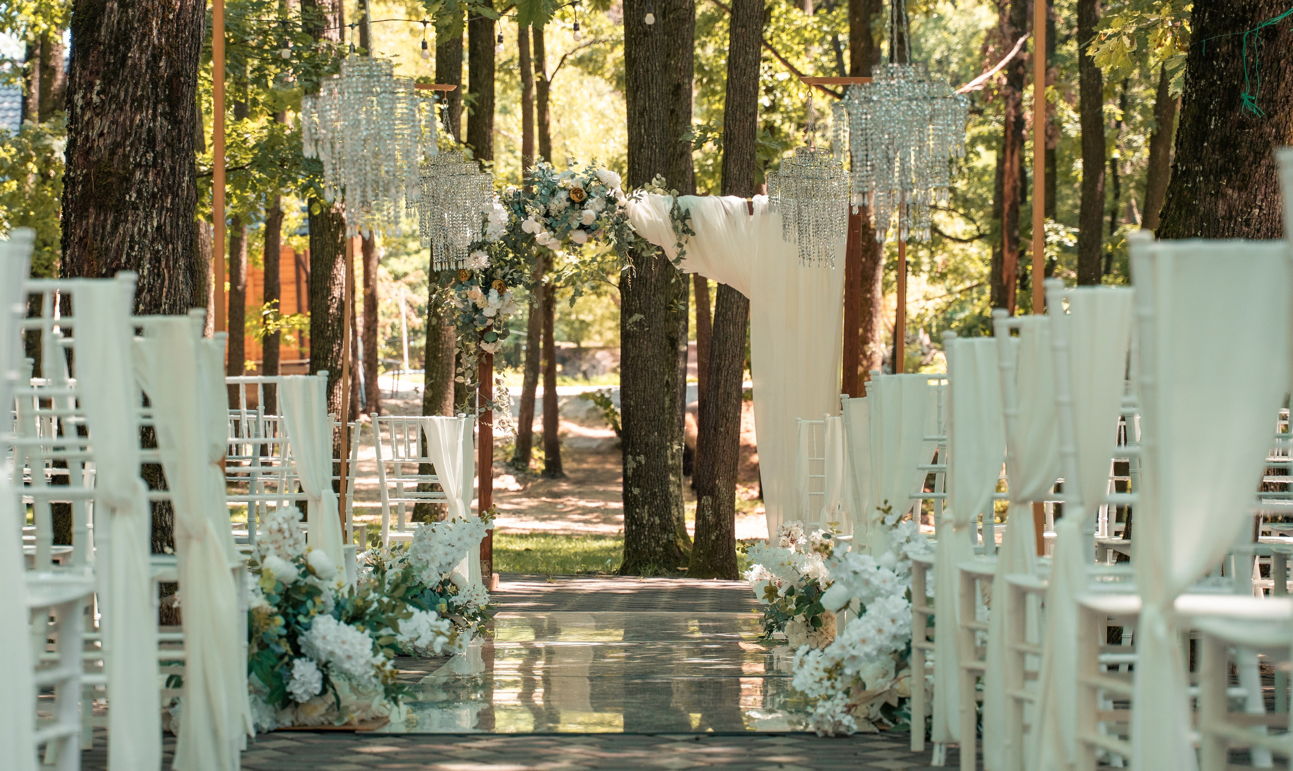Decor ceremonie nuntă în pădure, scaune albe cu voal, candelabre suspendate în copaci și arcadă florală, locație nuntă București, incadru.ro.