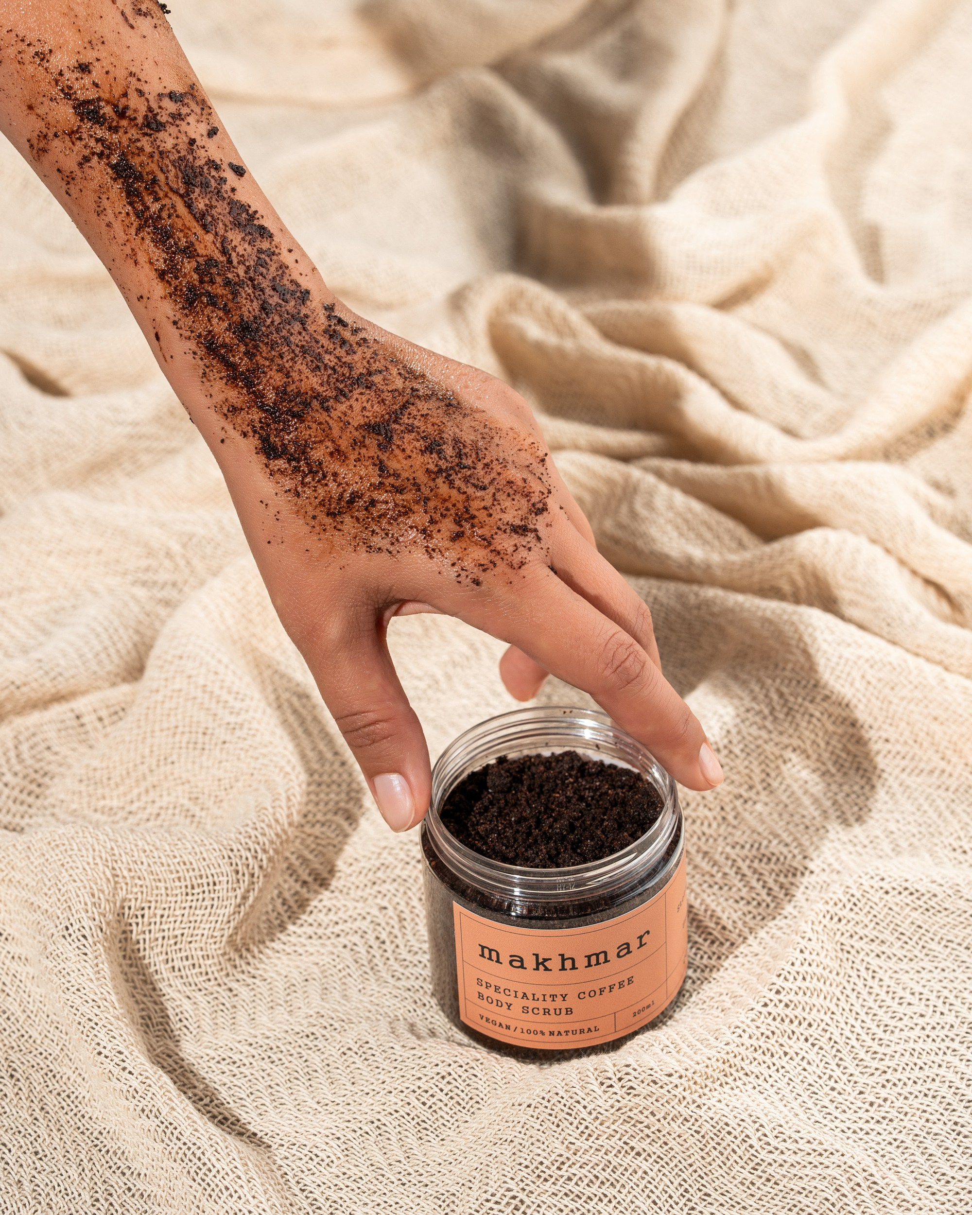 Makhmar Speciality Coffee Body Scrub açıq qabda, qəhvə skrabı ilə örtülmüş əllə bej toxumalı parça üzərində təqdimat.