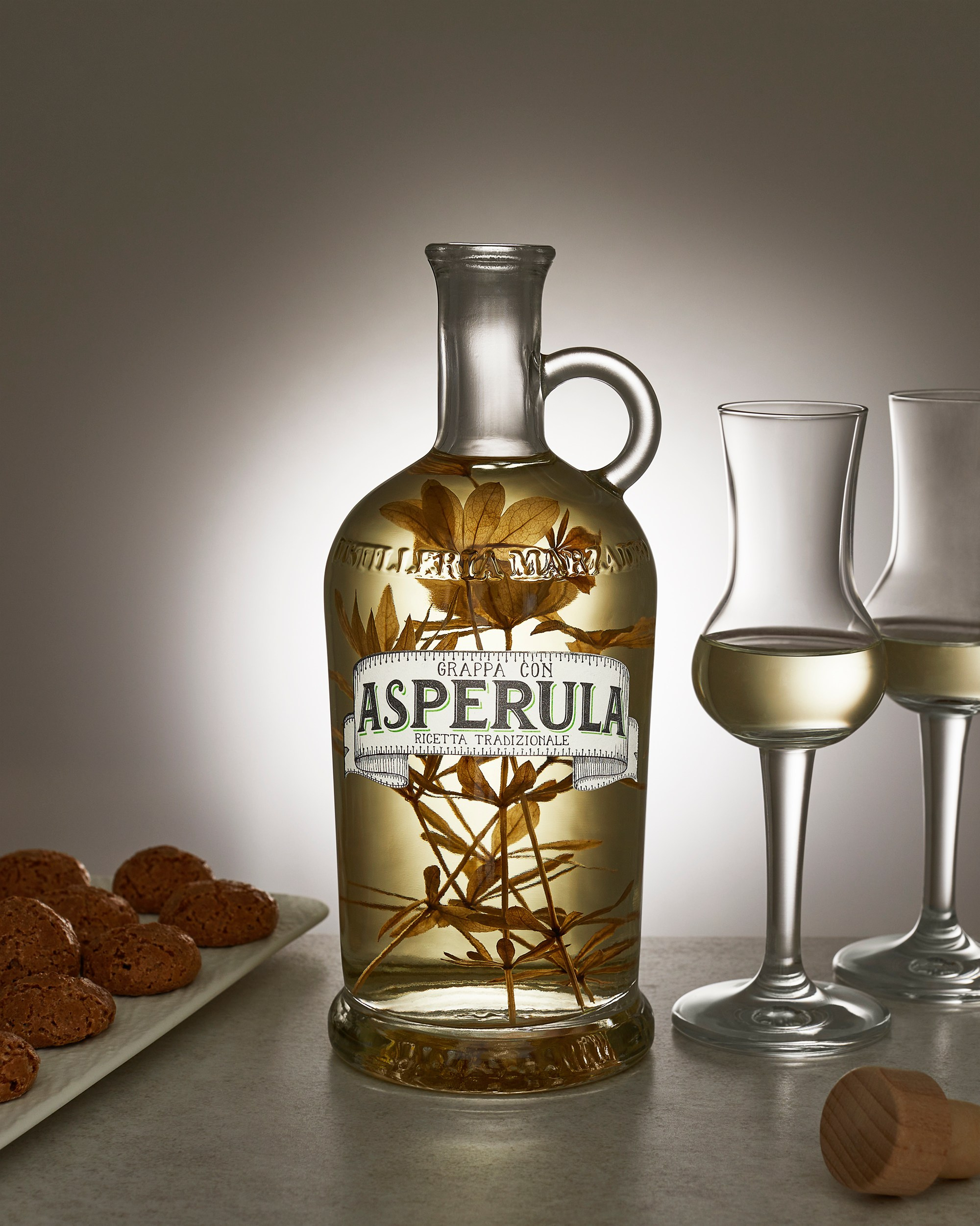 Photographer Roman Djuzev - A bottle of grappa with asperula grass, two glasses of grappa and a light snack of almond cookies on a stone table.