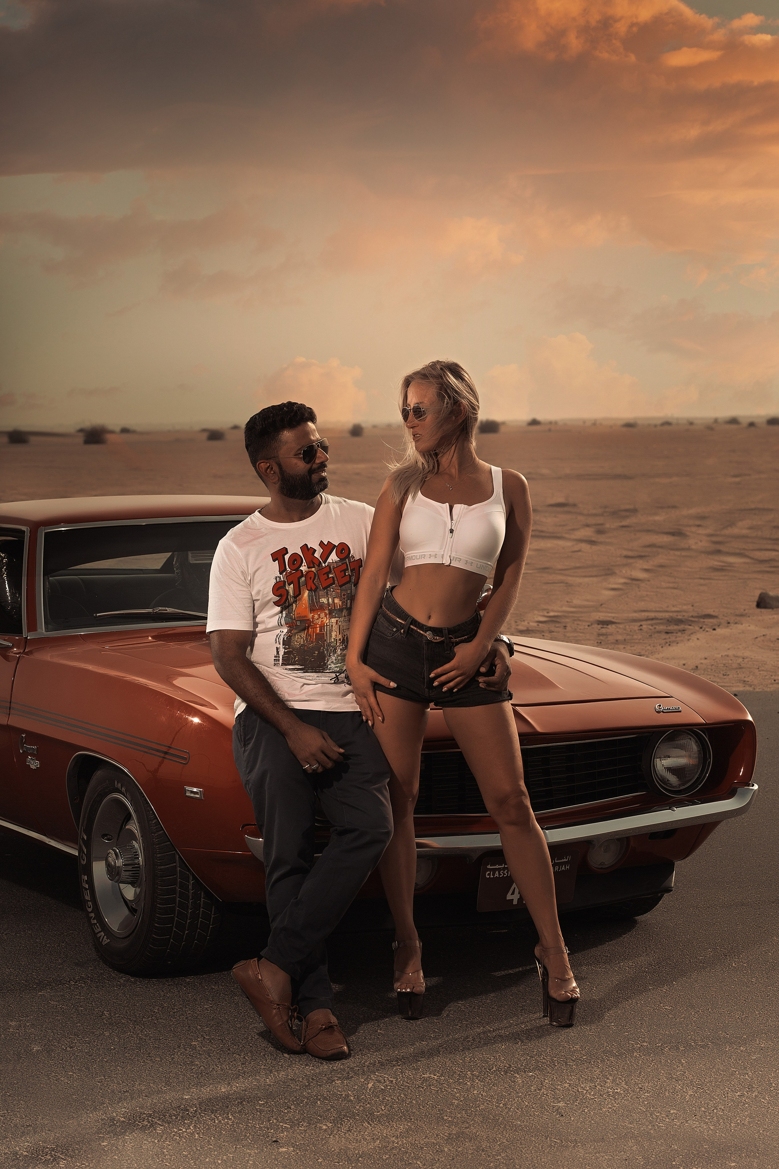 A beautiful woman sits with a guy on the bonnet of a stunning red car against the backdrop of Dubai’s sandy roads during sunset