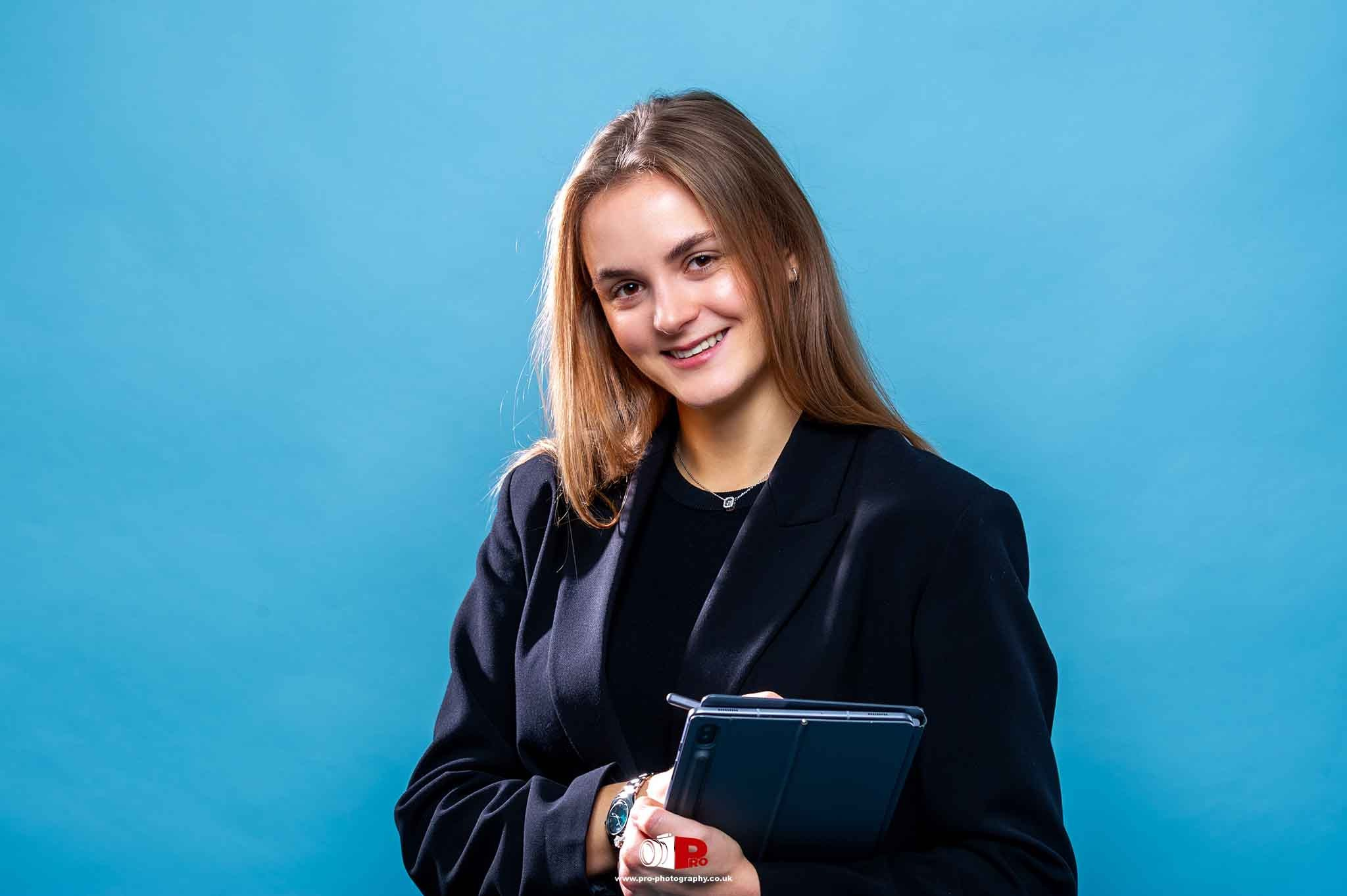 A young businesswoman in a navy blazer holding a tablet, posing confidently in front of a blue background.