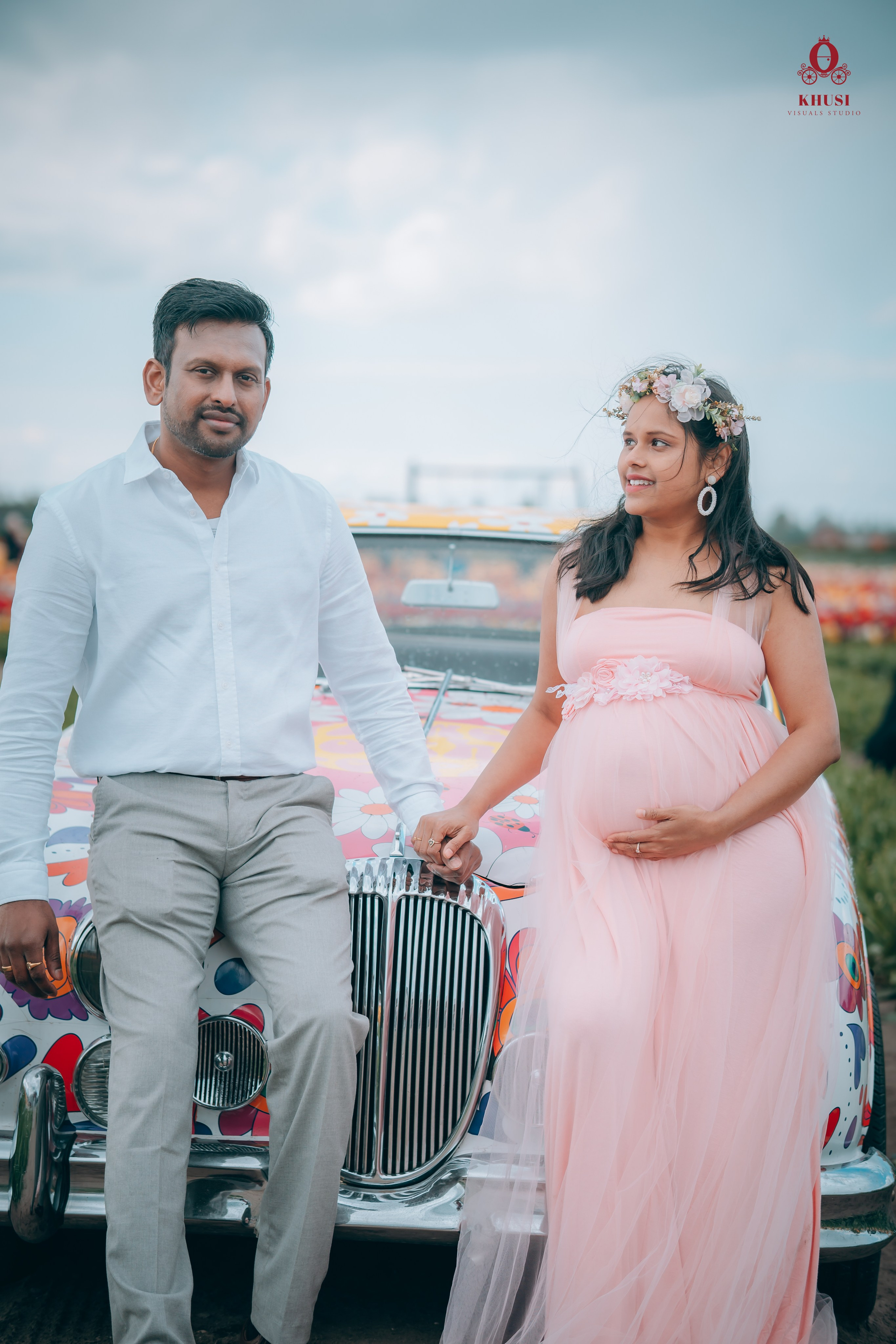 A pregnant woman holding hand of her husband and leaning on a car with floral print in a tulip field in Netherlands