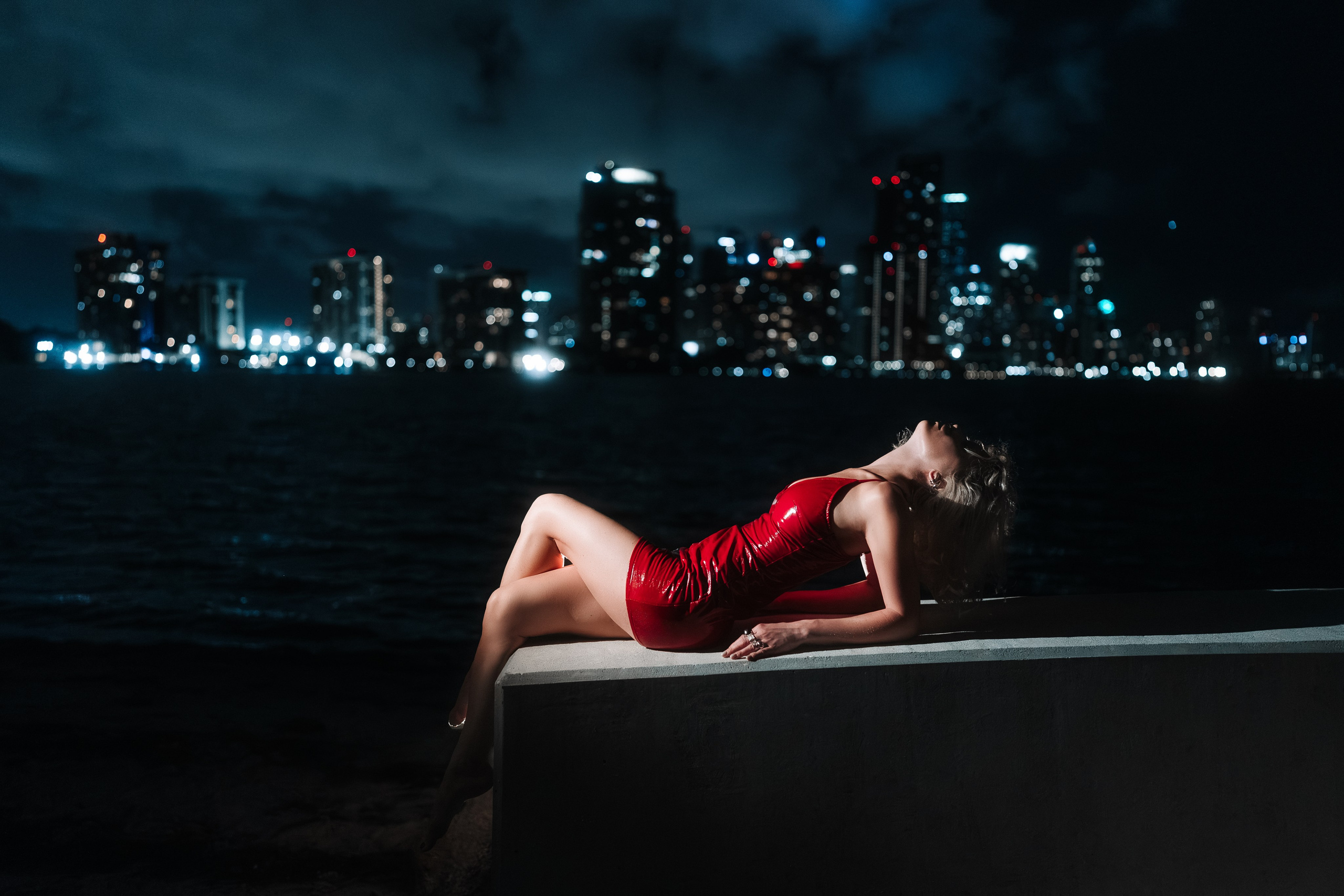 Full-body portrait by the water. Red dress, reflections across the bay, Miami skyline in the background. Hard directional lighting adds depth and dimension. Professional urban portrait session.