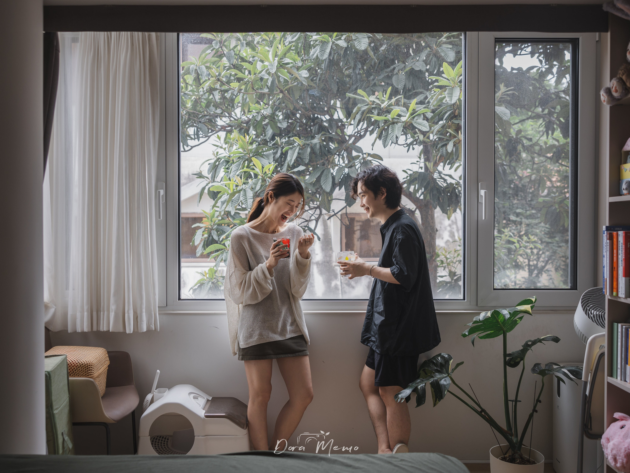 A couple laughs together in front of a large leafy window during a relaxed Shanghai lifestyle photography session.