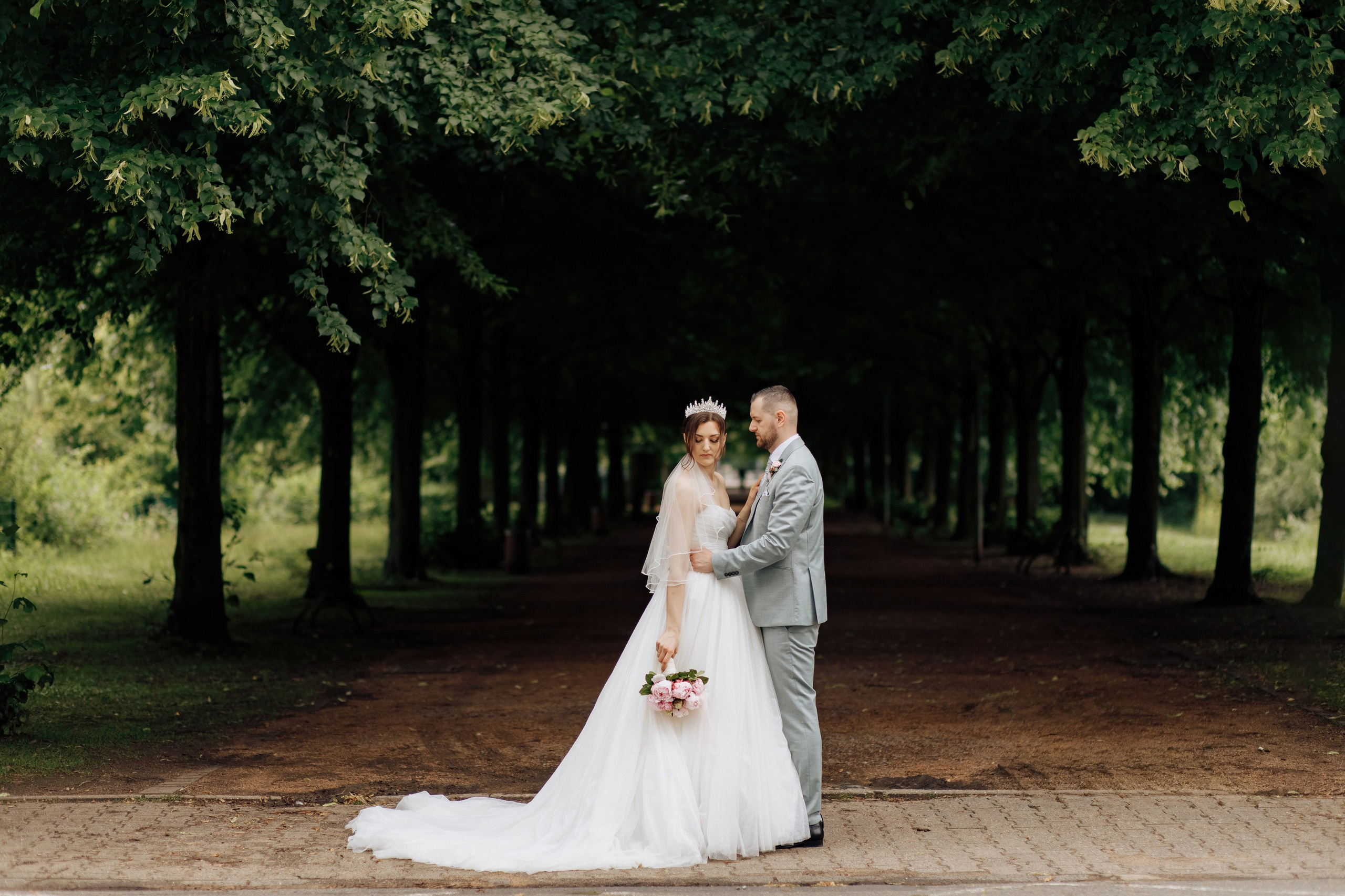 Der Bräutigam und die Braut stehen in einer Baumallee, während er sie verliebt anschaut. Ein romantischer Moment der Hochzeit.