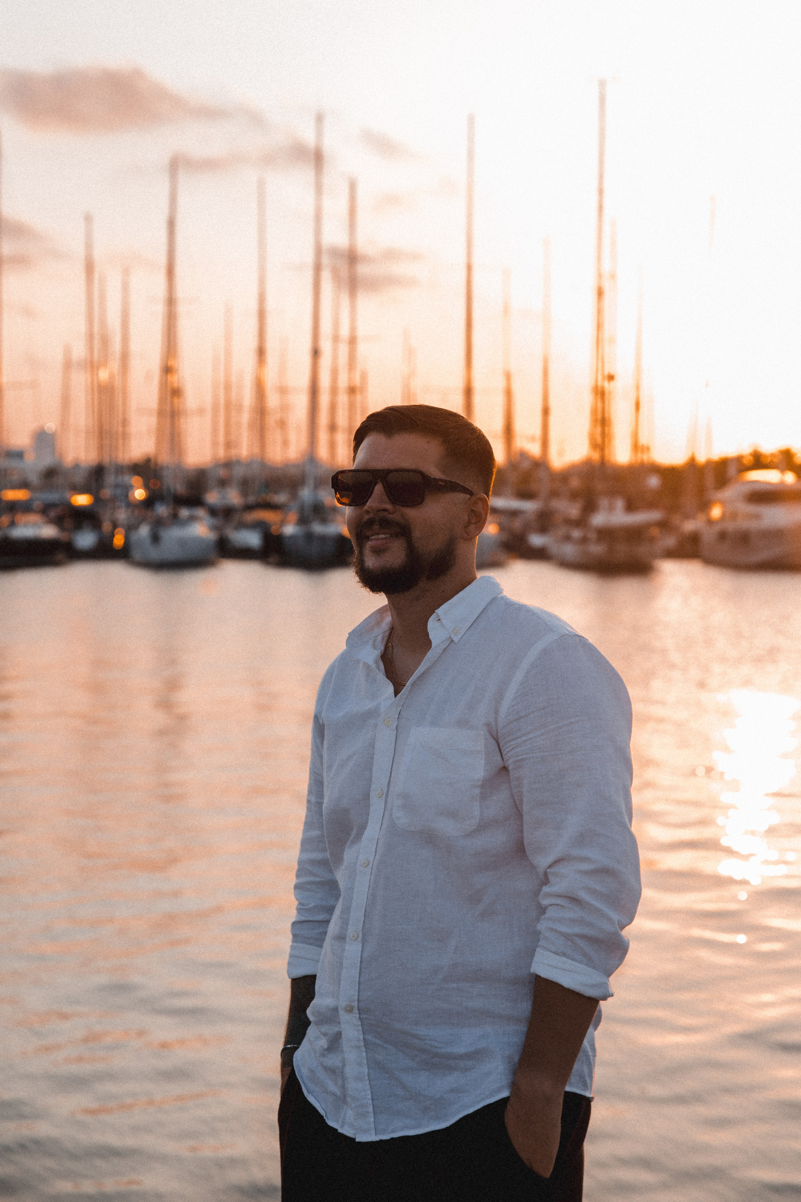 Мужская портретная фотосессия в Валенсии, Испания, с уверенным мужчиной в белой льняной рубашке и солнцезащитных очках у марины на закате, с парусными лодками и золотым средиземноморским светом на фоне, создающим расслабленное и стильное lifestyle-настроение — идеально для мужской портретной фотографии, lifestyle-фотосессий для мужчин, портретов для персонального бренда, casual fashion-сессий и профессиональных мужских фотосессий в Валенсии и по всей Испании.