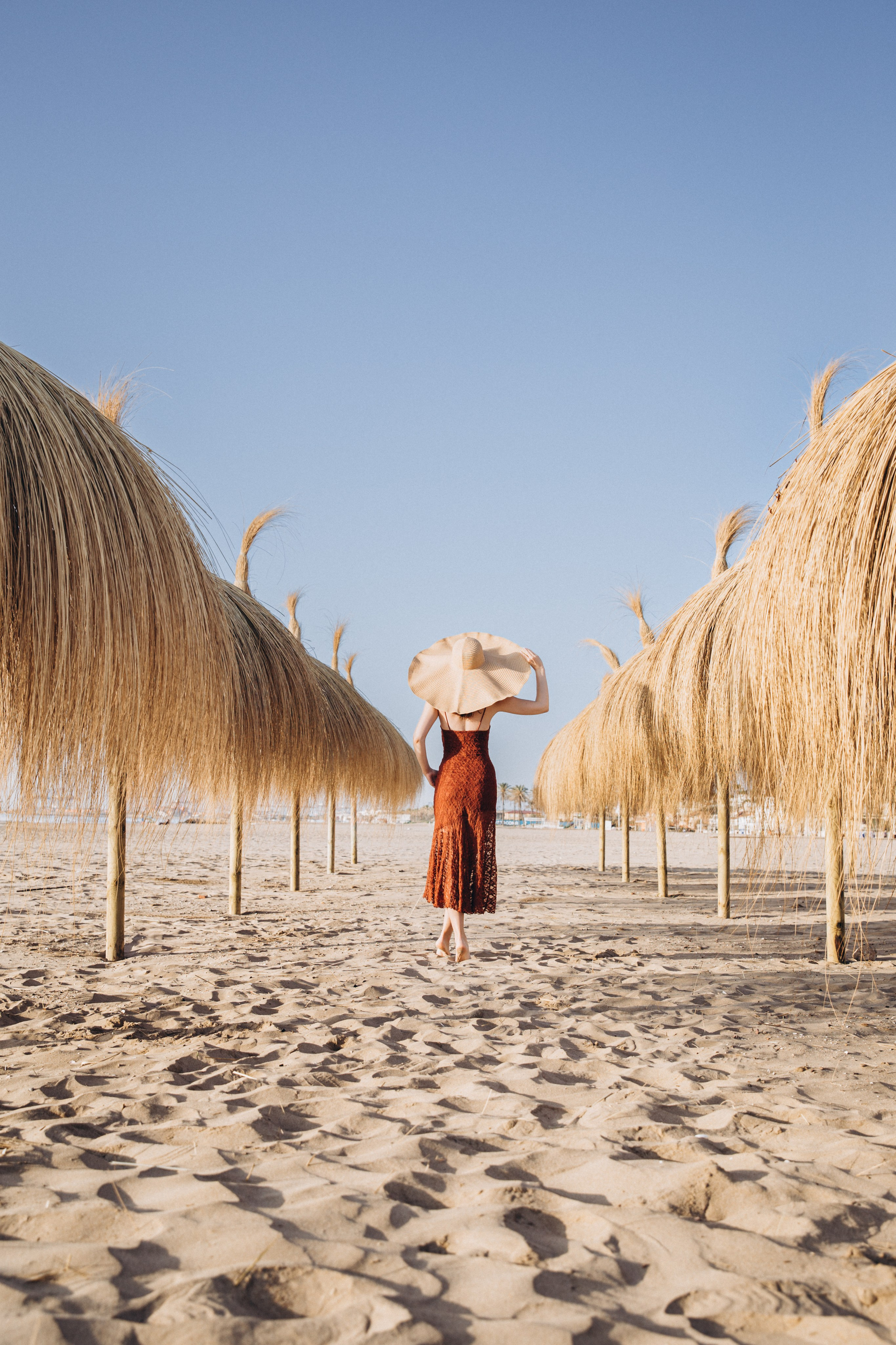 Женщина в шляпе с широкими полями идет босиком по песчаному пляжу среди соломенных зонтов в Кальпе, Испания — художественная летняя портретная фотосессия, отражающая расслабленную атмосферу побережья и красоту Средиземноморья.