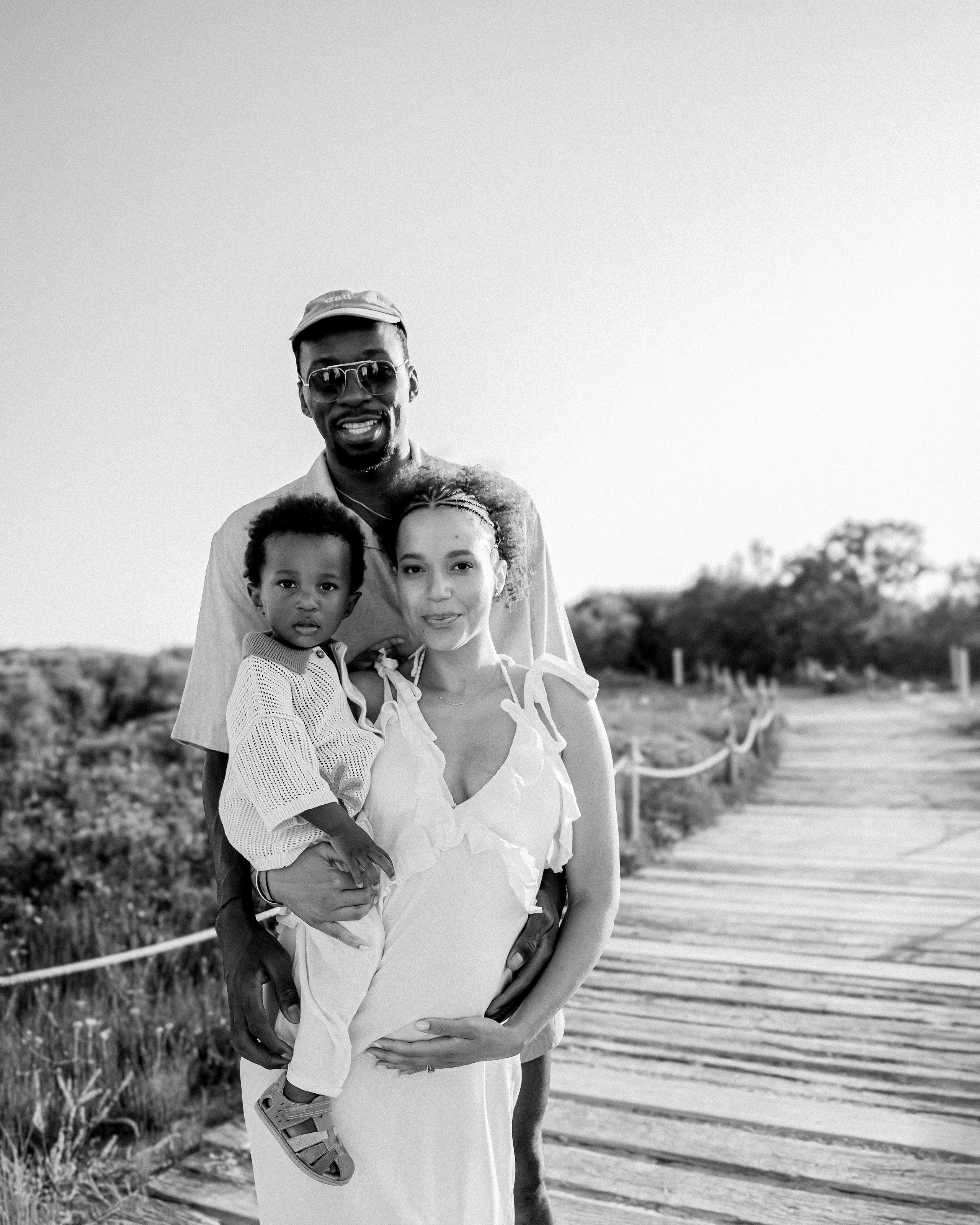 Retrato familiar en blanco y negro sobre una pasarela de madera al atardecer en Alicante, España — pareja embarazada feliz con su hijo pequeño, ideal para sesiones elegantes y emocionales en la Costa Blanca.
