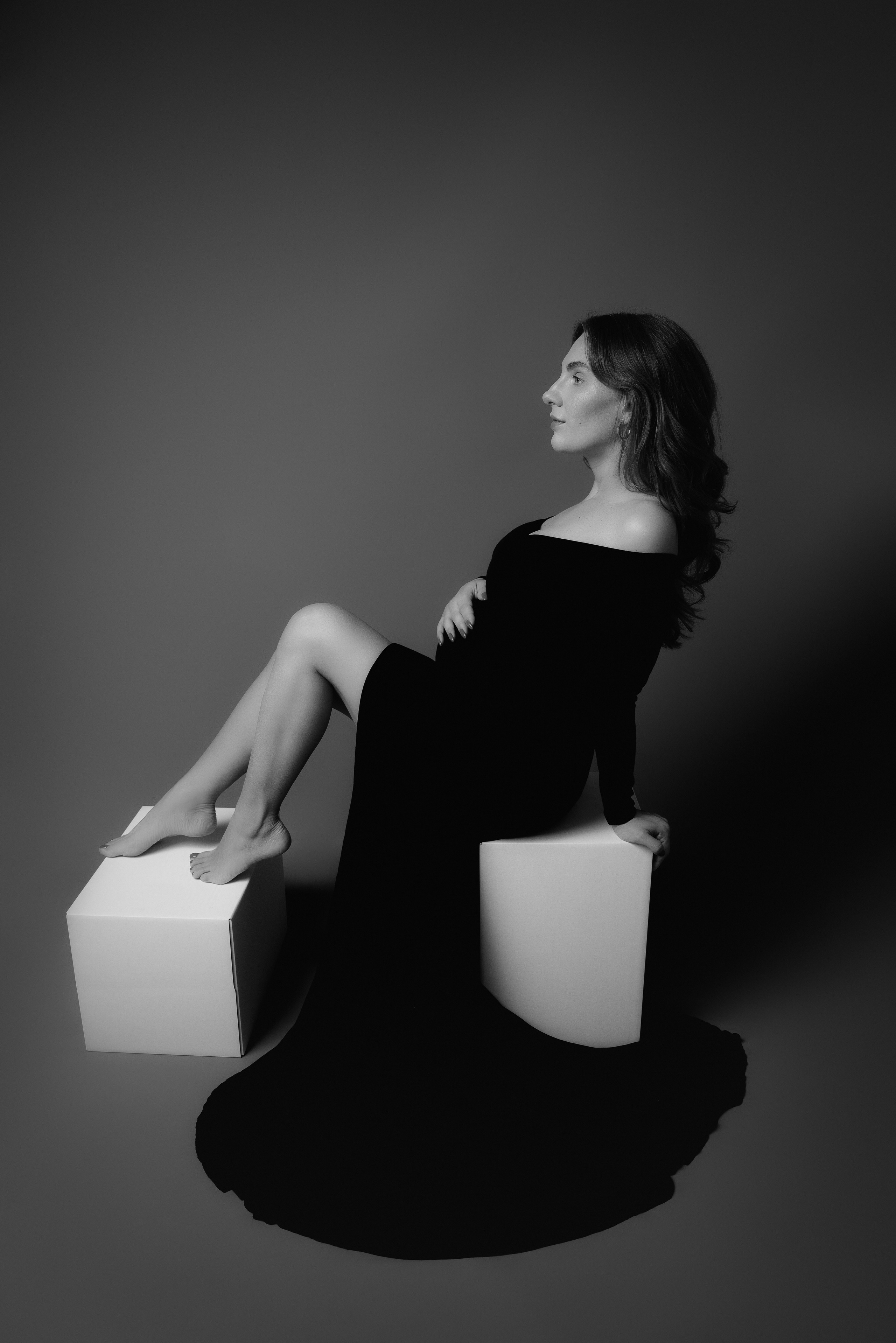 schwangere Frau posiert im Studio in einem schwarzen Kleid auf einem weißen Würfel