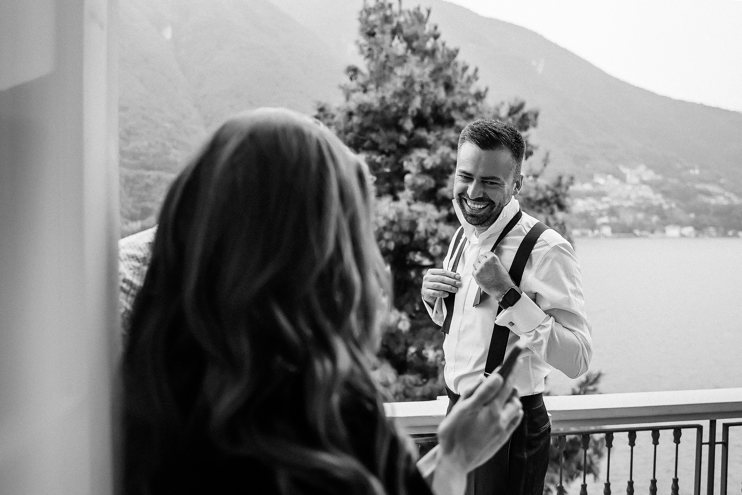 Black and white photo of a groom adjusting suspenders on a balcony, with mountains and a lake in the background. The bride stands nearby with long hair, capturing a candid pre-event moment amidst scenic beauty.