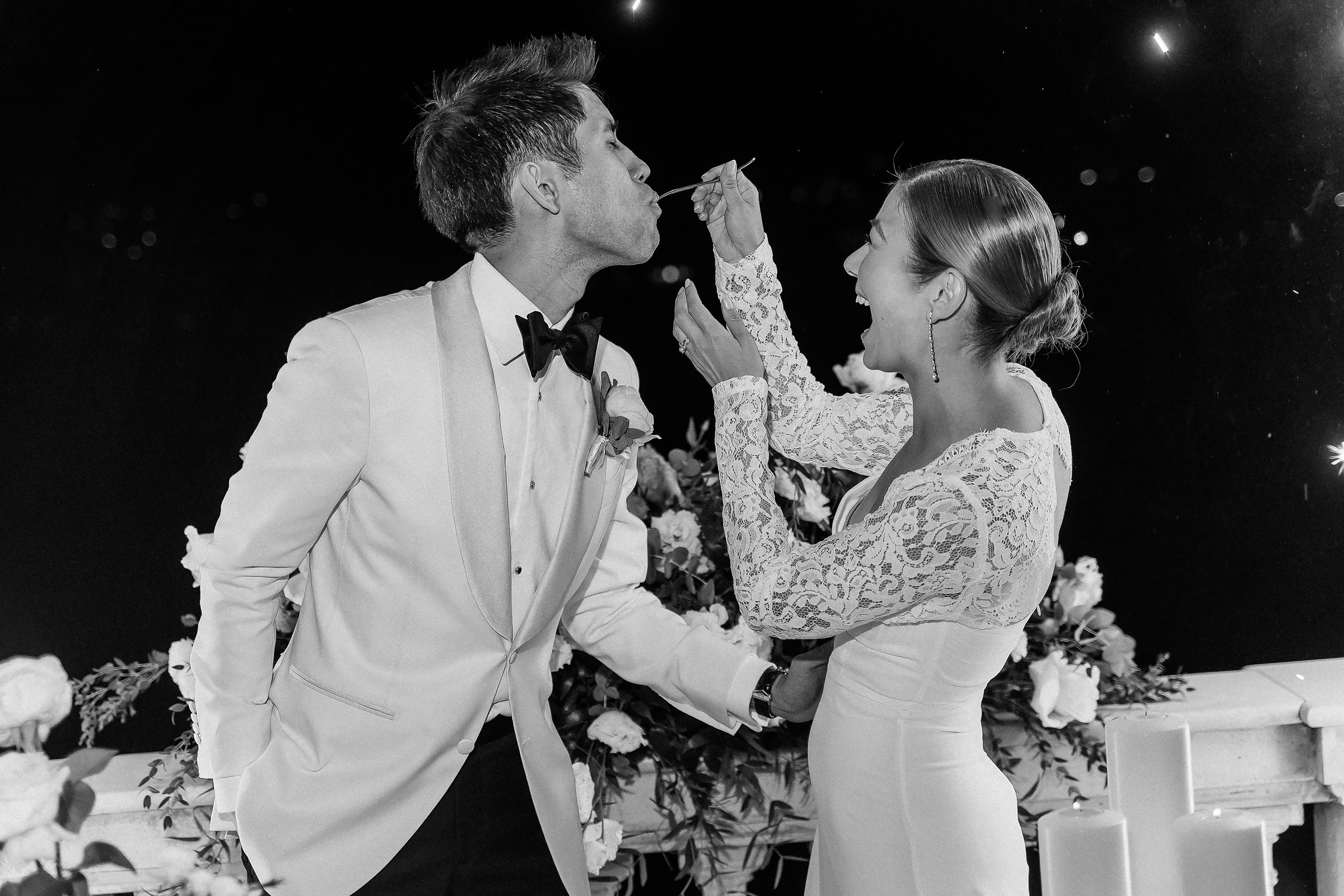 Groom leans forward to accept a bite of cake from the bride in a playful moment.