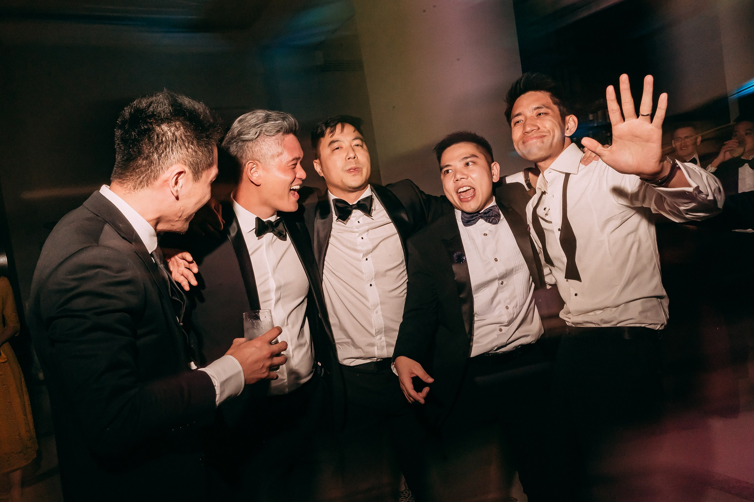 Group of five men in tuxedos with open collars, embracing and smiling widely on the dance floor.