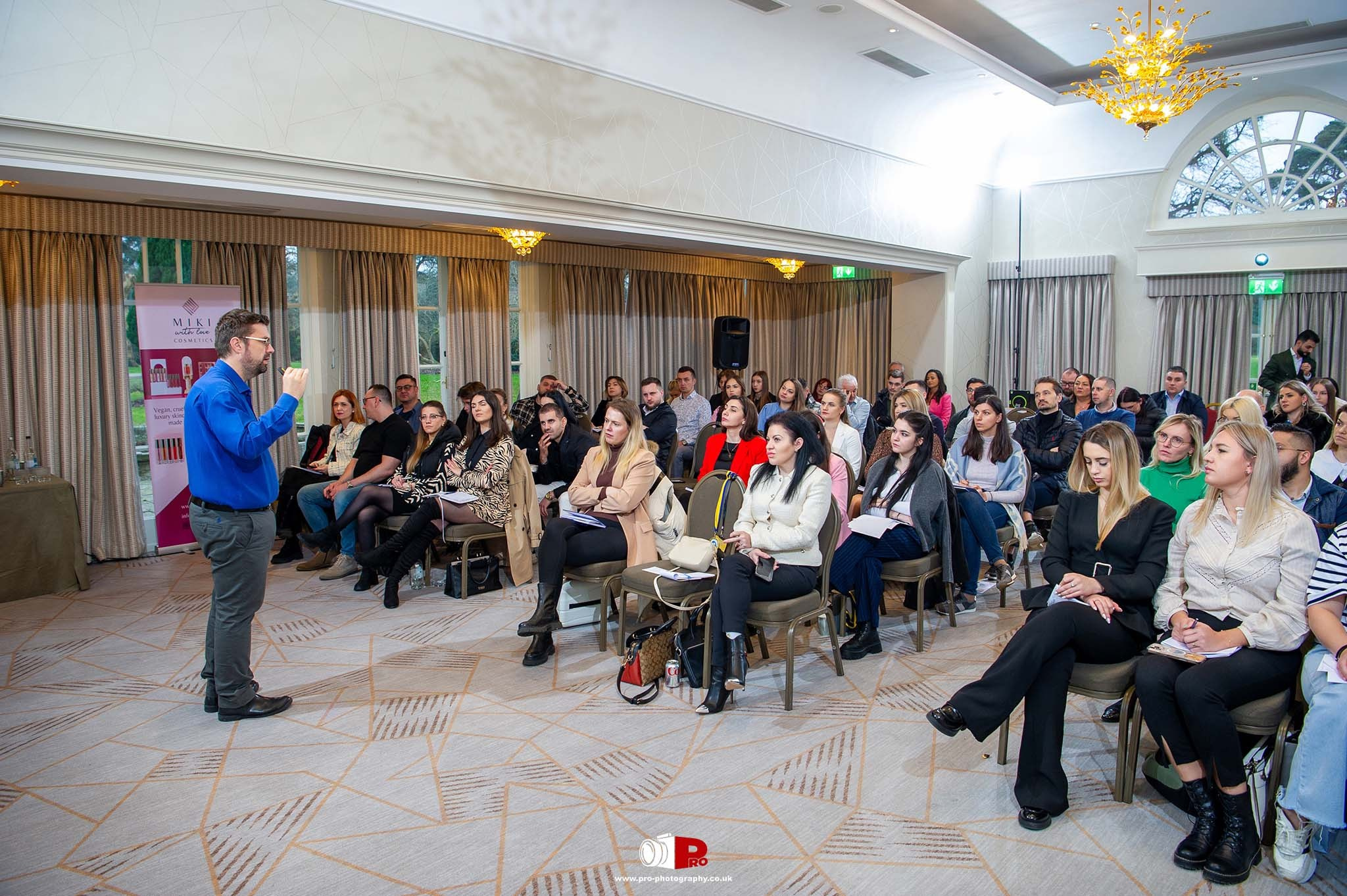 A room full of attendees listening to a speaker in a blue shirt presenting at a business conference.