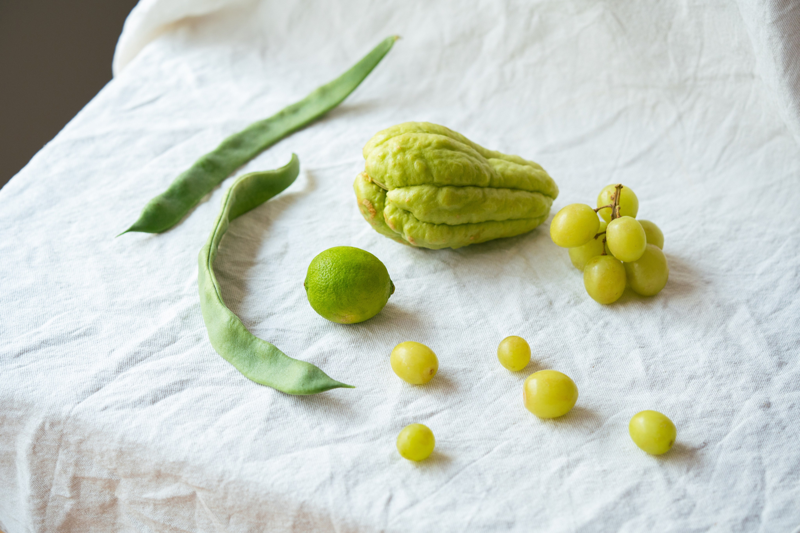 Minimalistic Food Arrangement with Grapes, Chayote, Lime, and Beans. Flat lay of fresh produce including green grapes, chayote, lime, and green beans on a white textured surface.