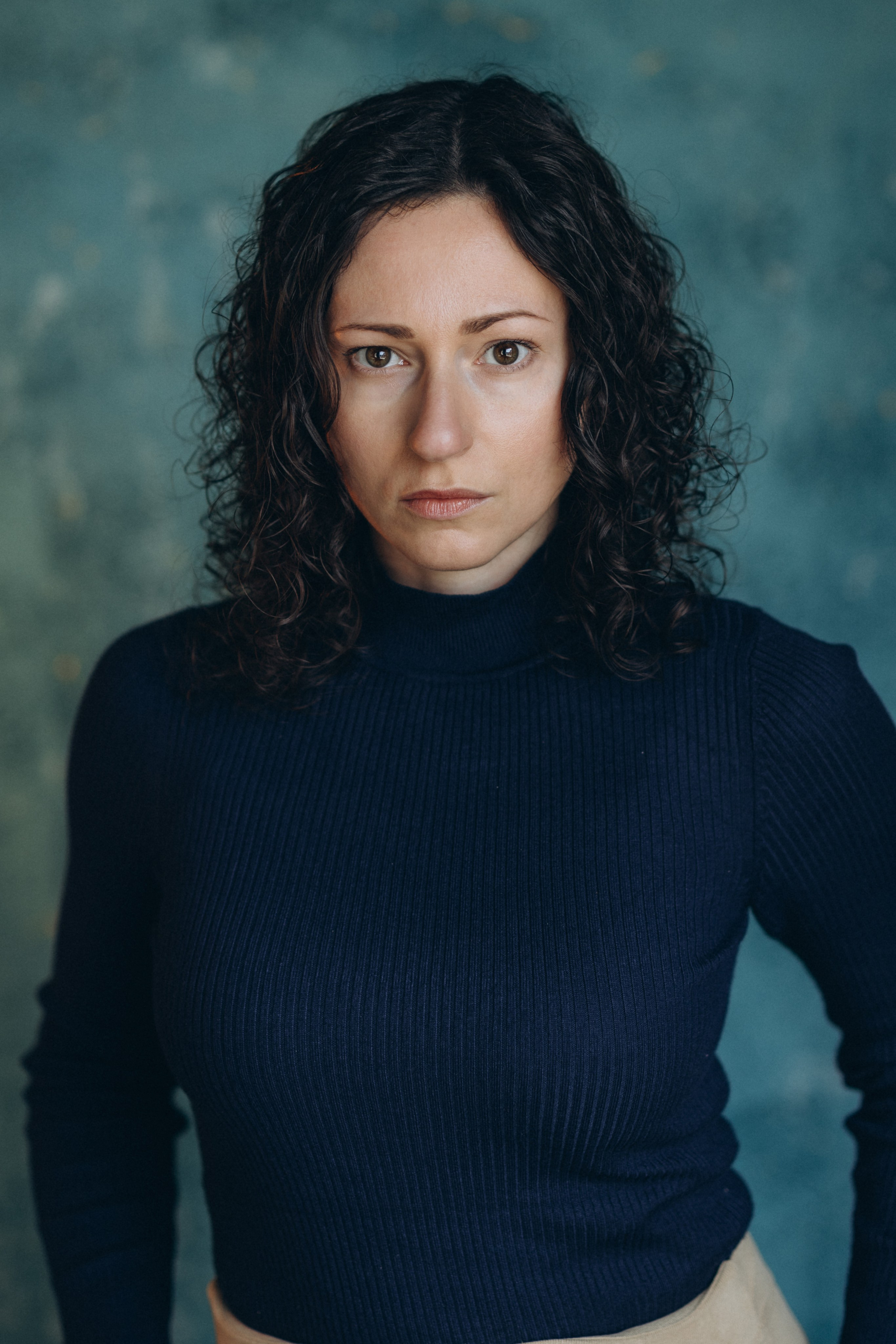 Actor headshot of a woman with curly hair in a navy blue top, looking into the camera with a neutral expression