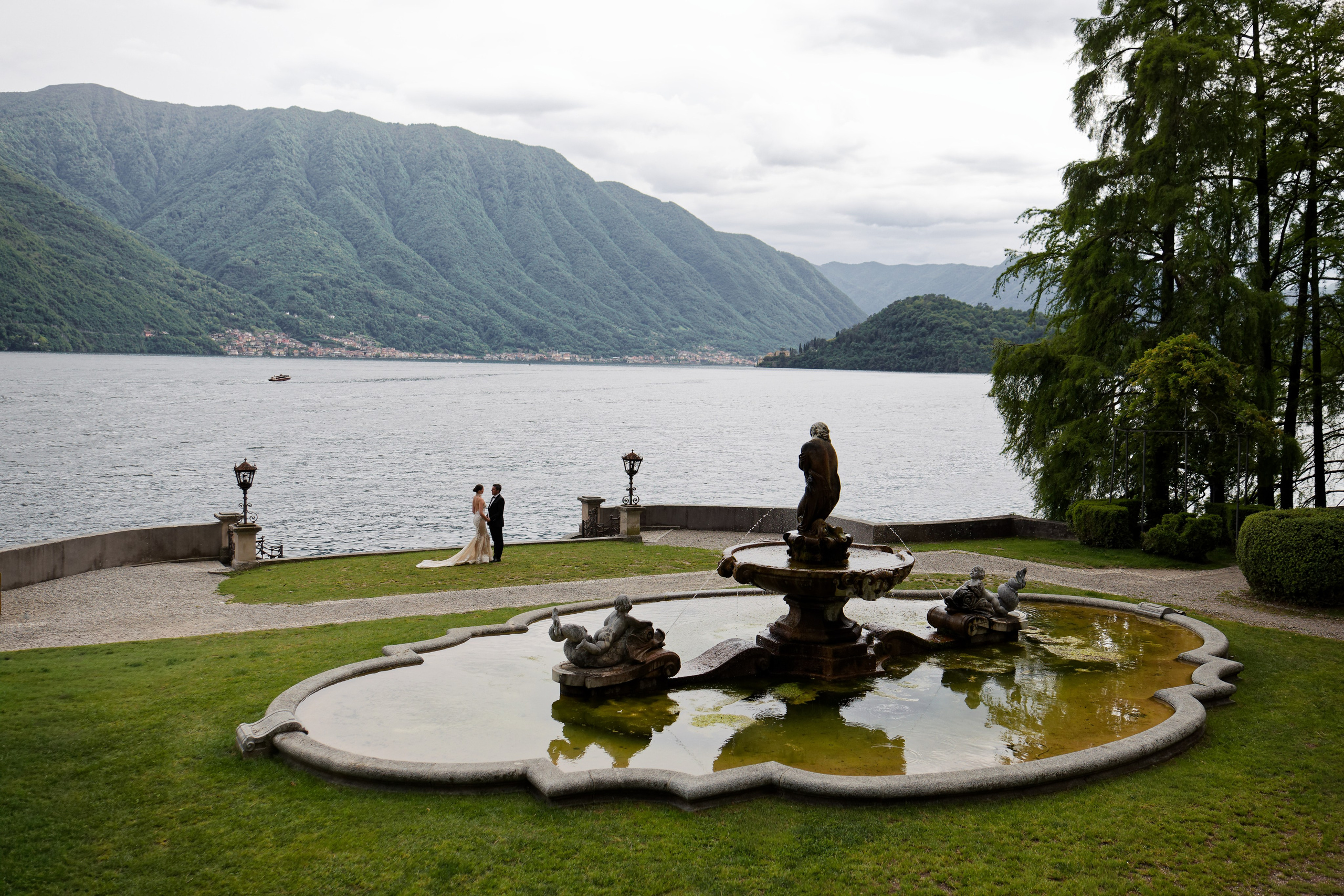 "Panoramic view of a lake and mountains with a fountain and park in the foreground."