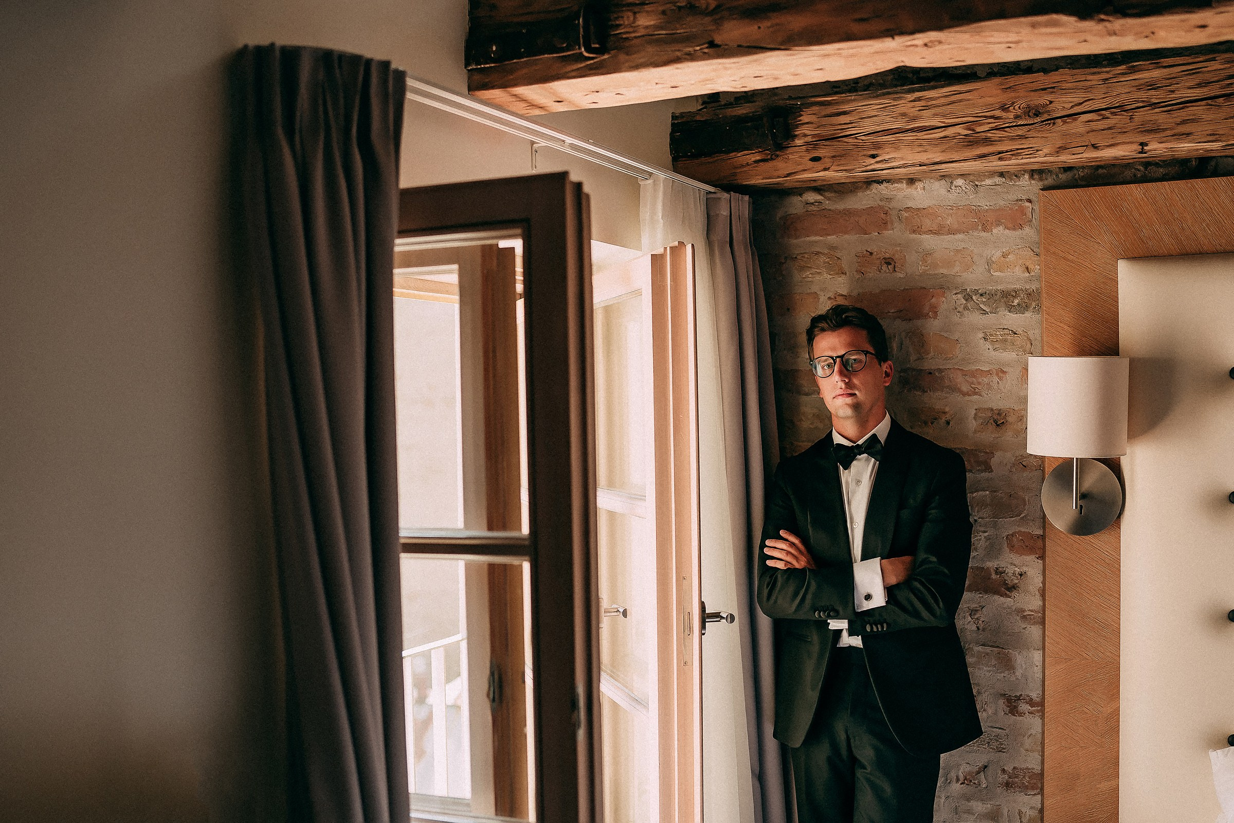 The groom dressed in a black tuxedo with a white shirt and black bow tie is standing with arms crossed next to a window. The background features a brick wall and a wooden beam ceiling